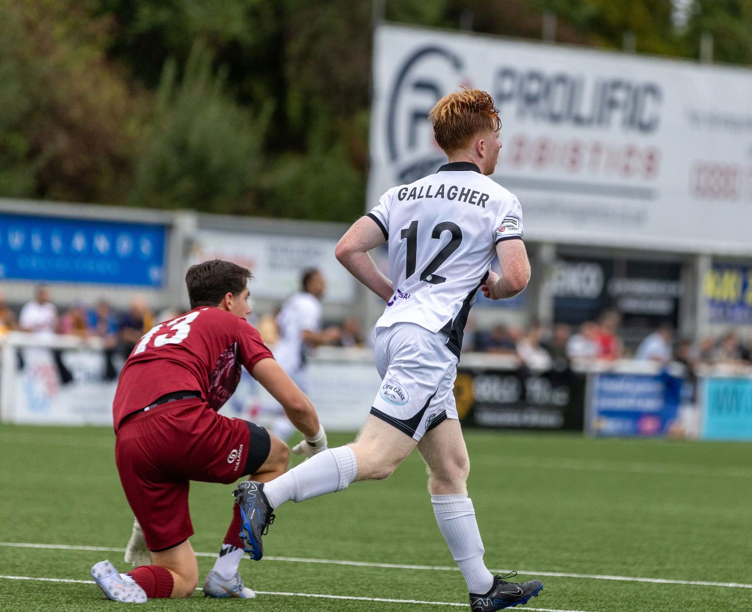 Soccer player Gallagher wearing number 12 white jersey dribbles past sliding opponent during outdoor match.