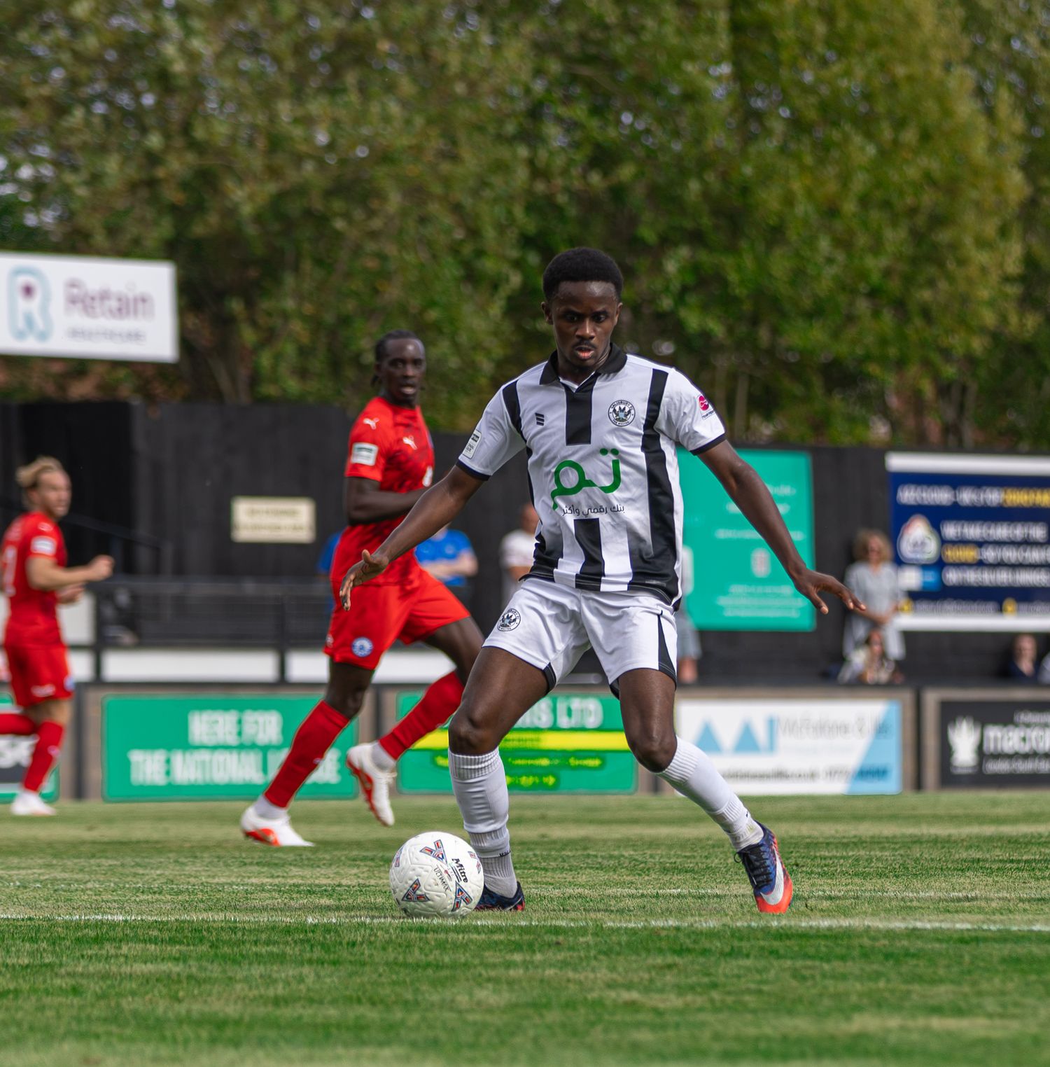 A young footballer in black and white kit dribbles the ball during a match with red-kitted players in background.