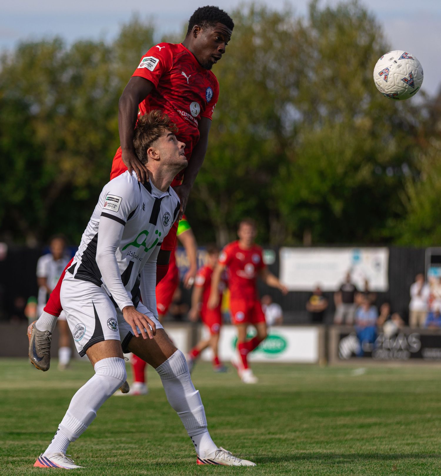Two soccer players compete for a header during a match, one in red and one in white and black uniforms.