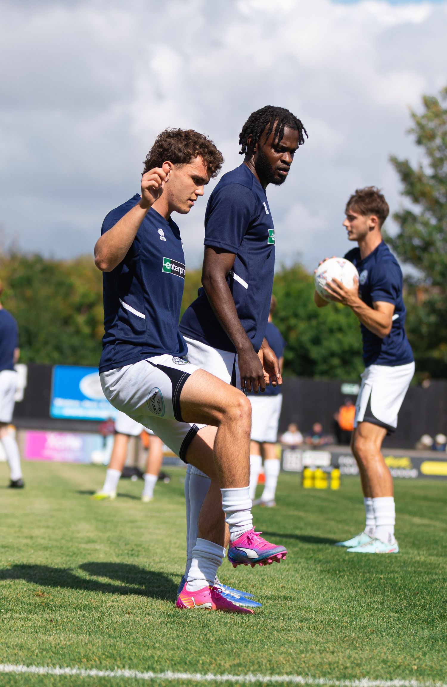 Soccer players in navy uniforms warming up on a grass field, one stretching with a raised knee during pre-match preparation.
