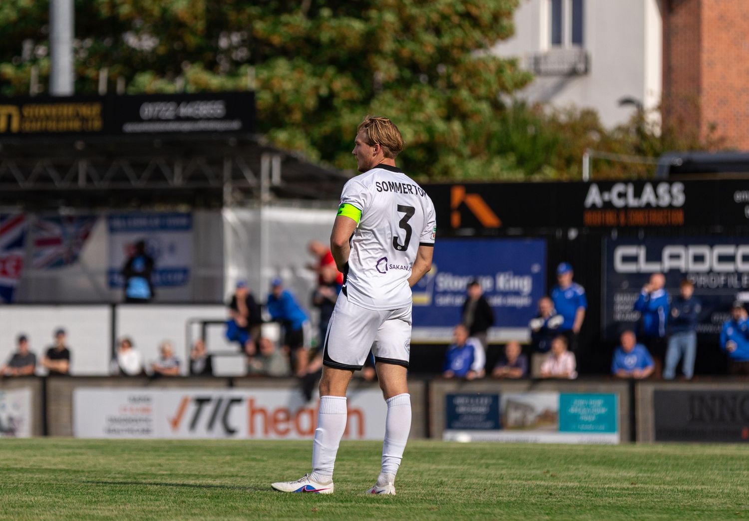 Soccer player wearing number 3 jersey stands on green pitch during sunny outdoor match with stadium signage visible.