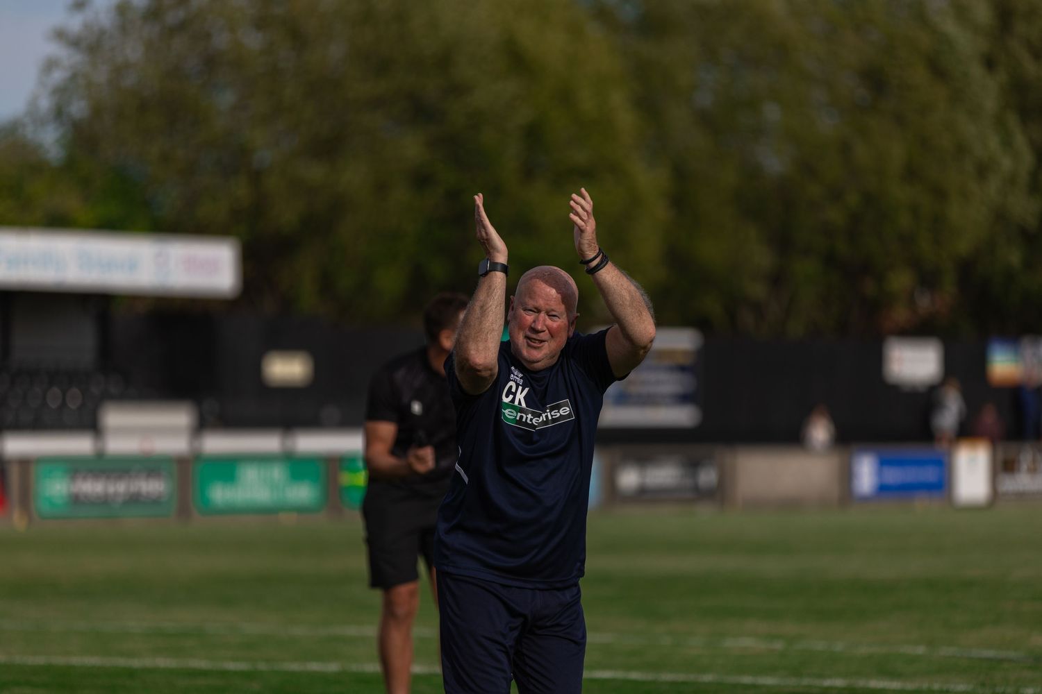 A football manager applauds fans on a sunny green pitch during a non-league match.