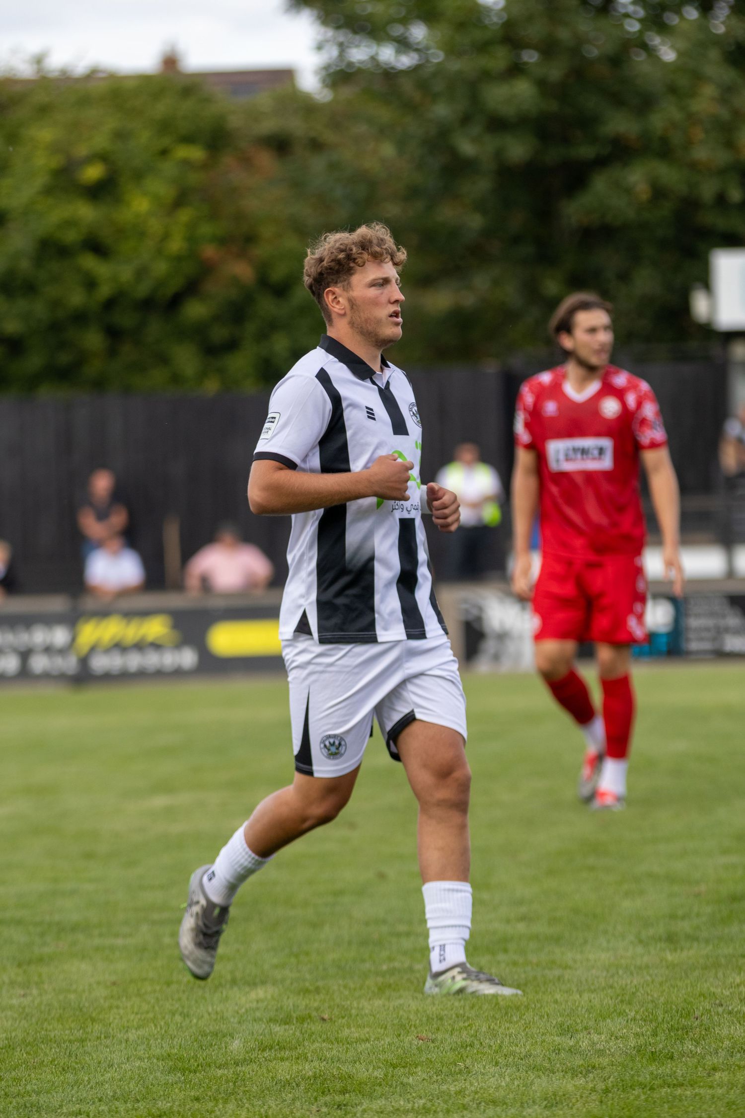 A male soccer player in black and white striped kit jogs on a grass pitch during a match, with an opponent in red visible behind.