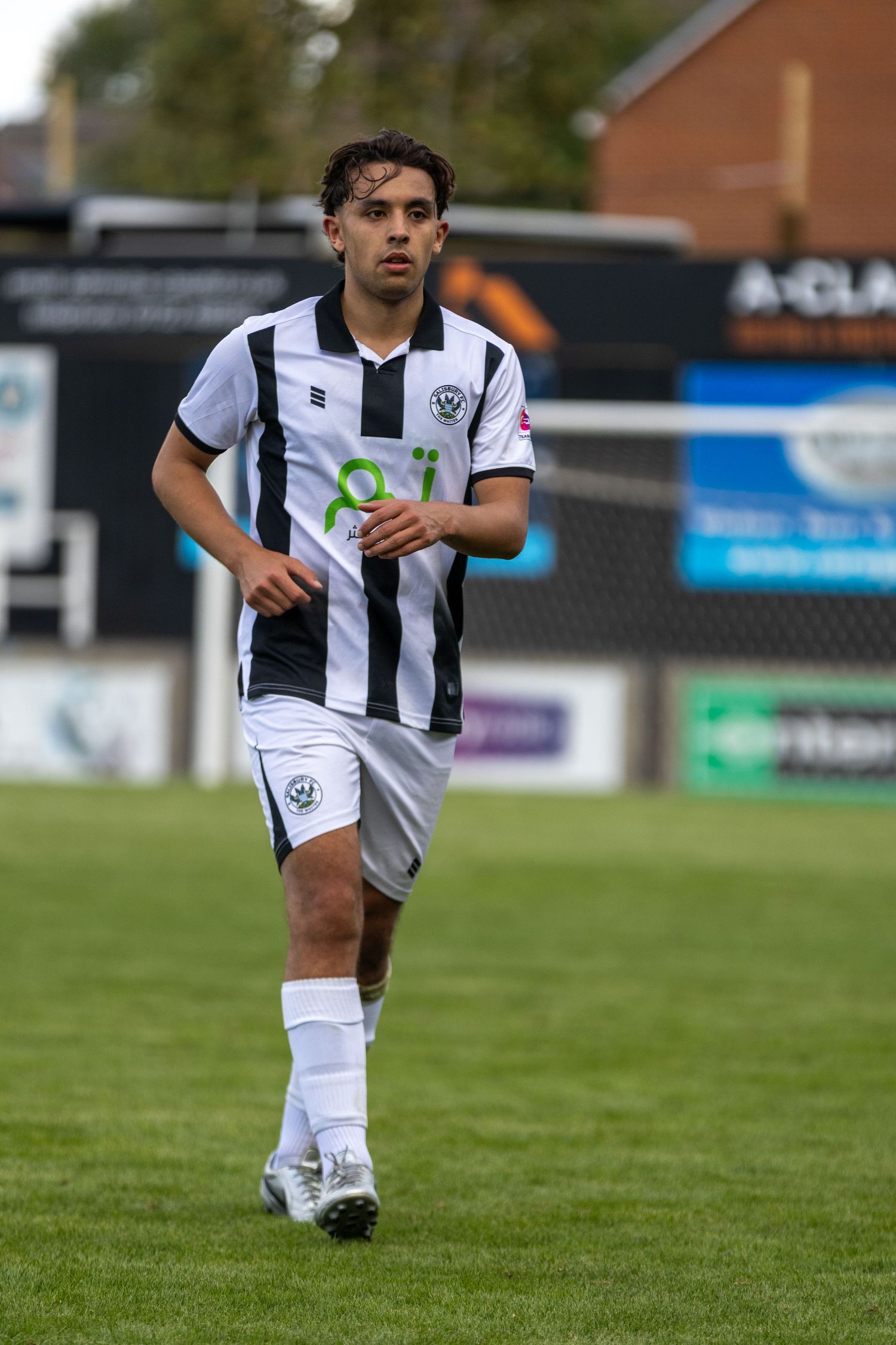 Young footballer in black and white striped kit jogging on green grass pitch during a match.