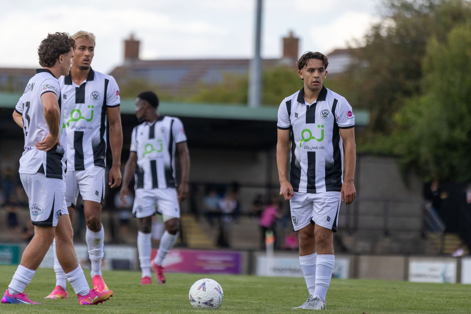 Soccer players in black and white jerseys stand near the ball during a free kick setup.