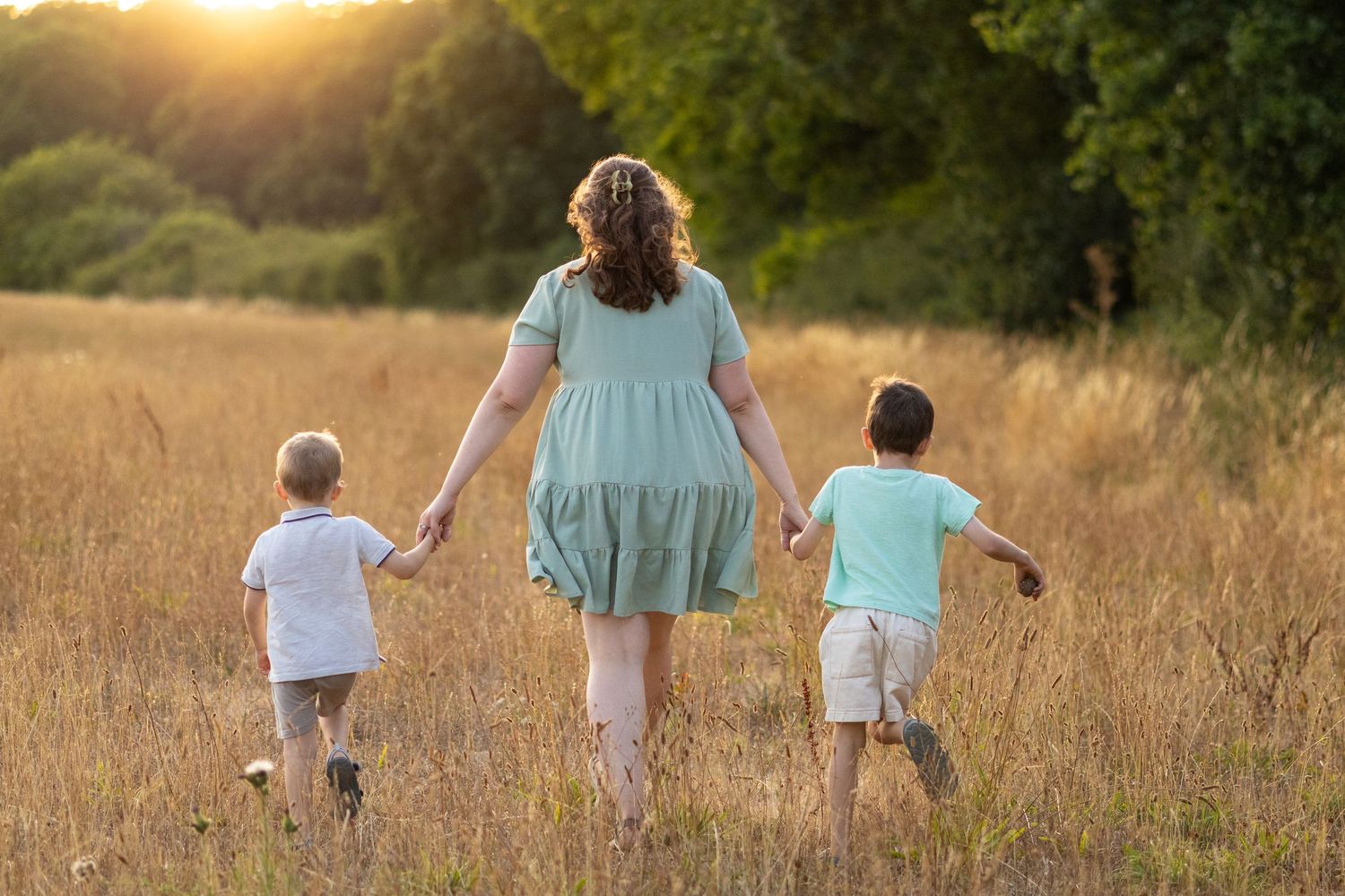 Mother and two sons walk hand-in-hand through dry grass field during warm summer sunset.