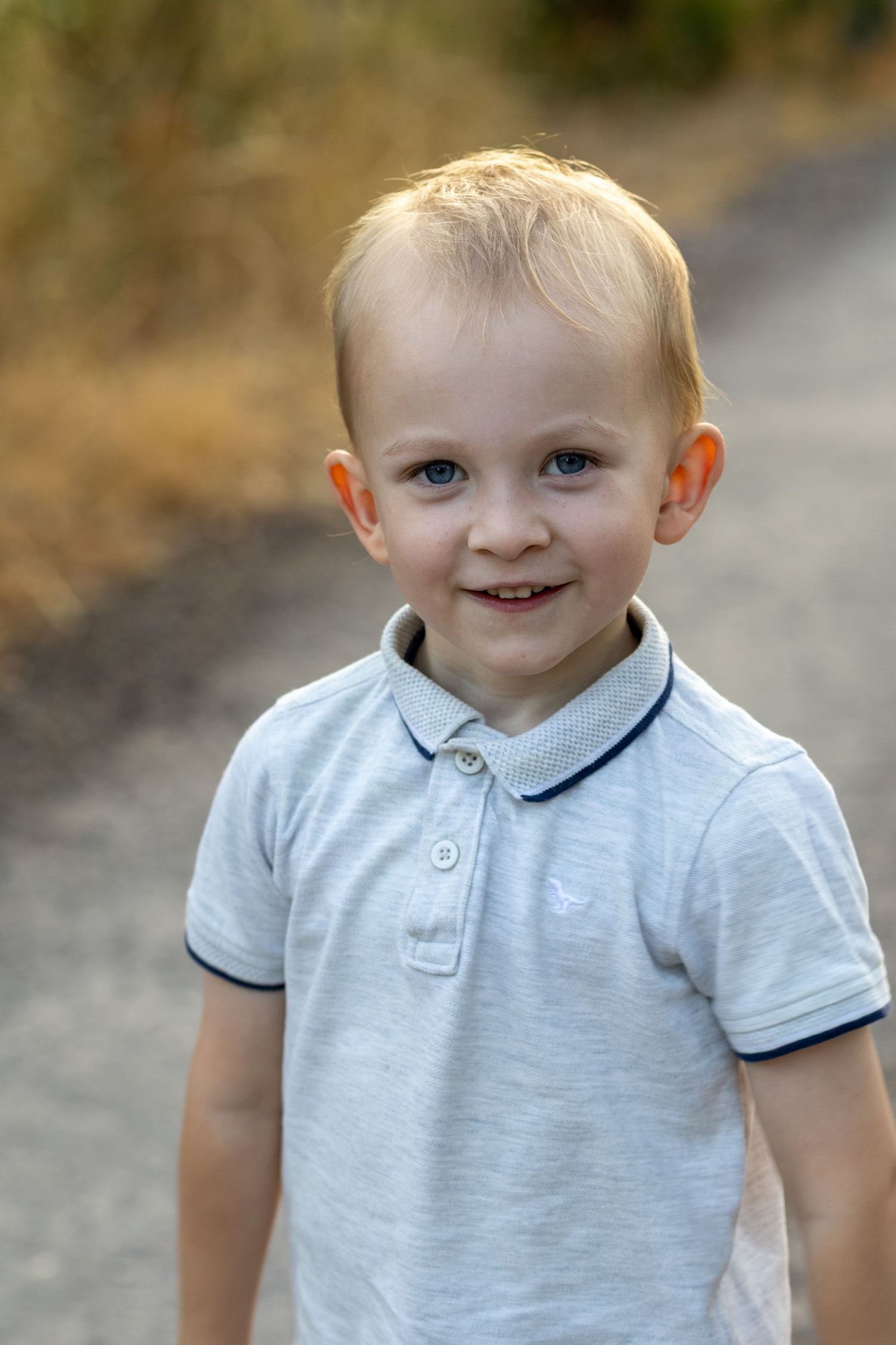 A cheerful blonde toddler boy in a grey polo shirt smiles on a sunny outdoor path.