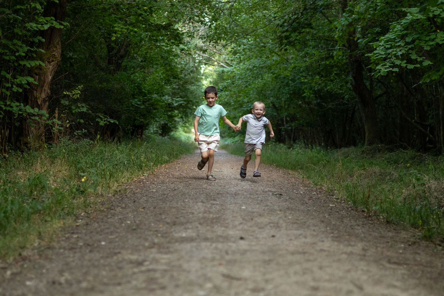 Two children run hand-in-hand down a sunlit tree-lined forest path.