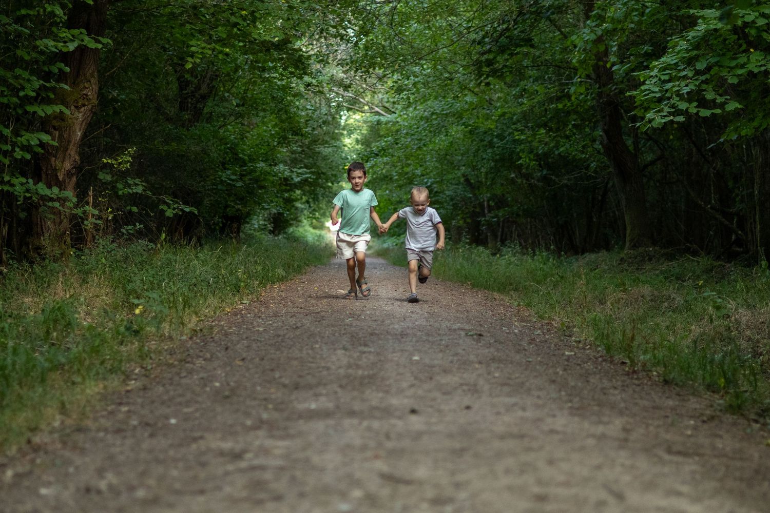 Two kids holding hands running together down a sunlit dirt path through a green forest.