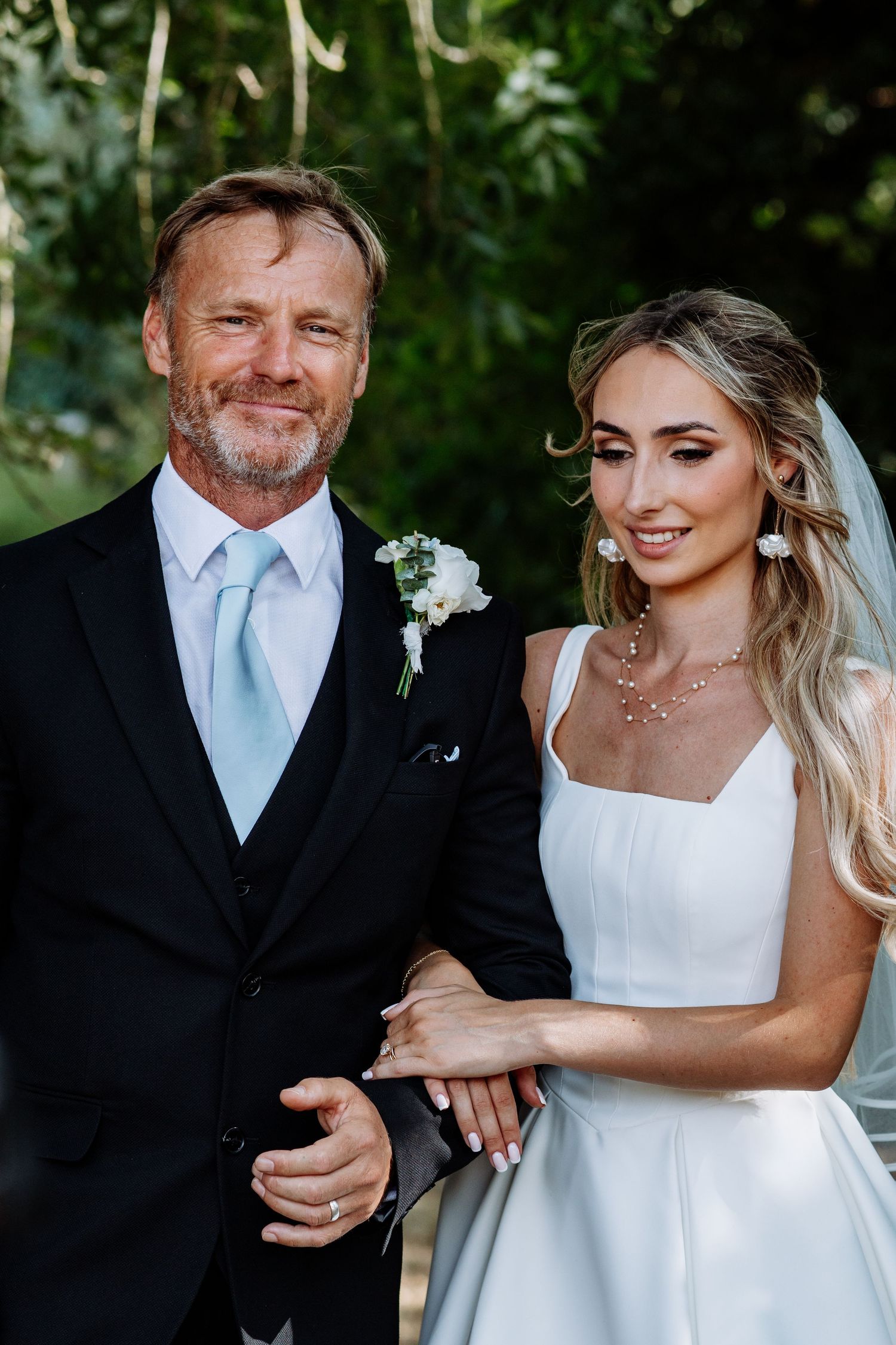 Bride in white gown smiling alongside older gentleman in black three-piece suit at outdoor wedding.