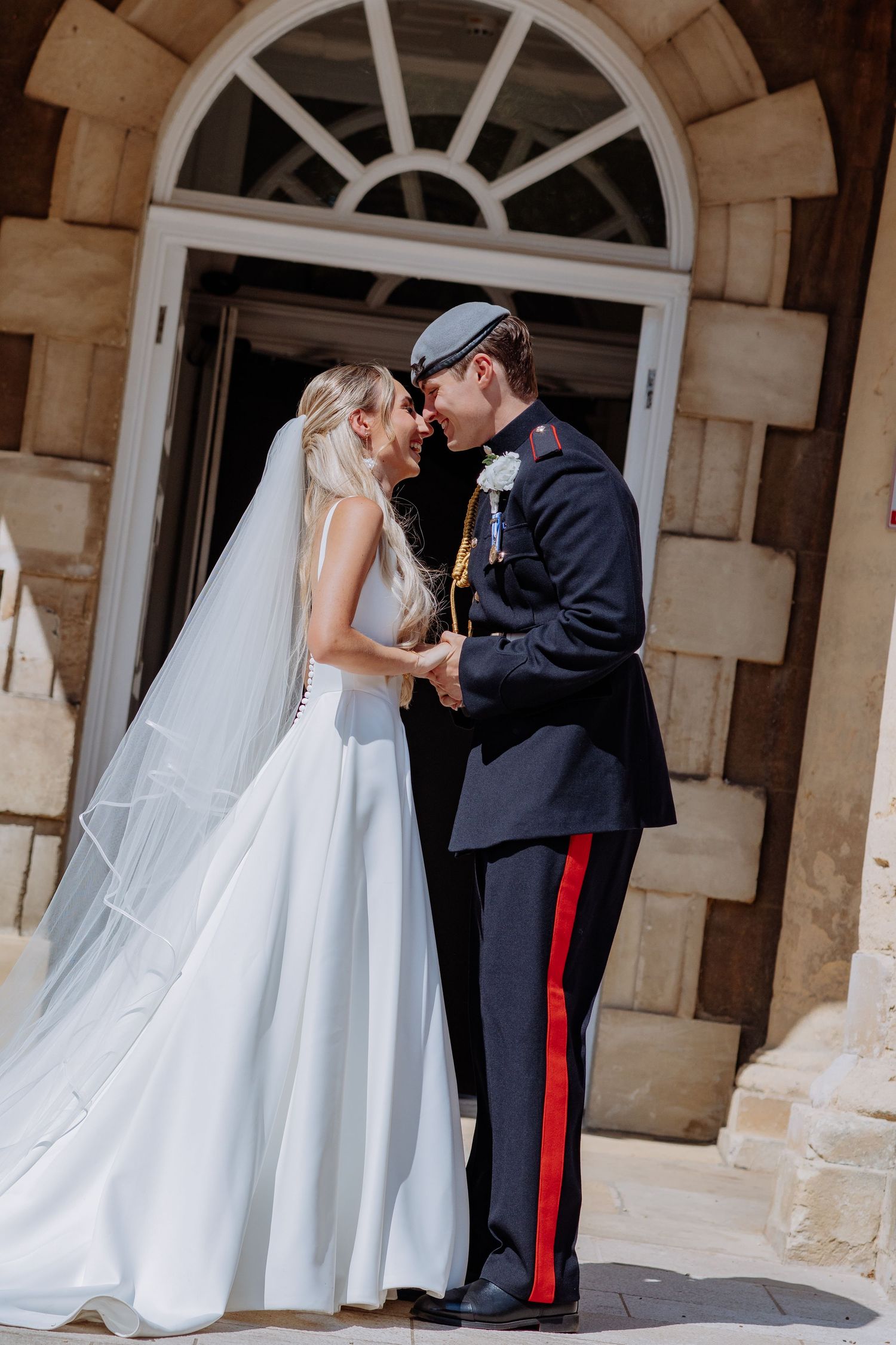Bride in elegant white gown and groom in military dress uniform share a tender moment outdoors.
