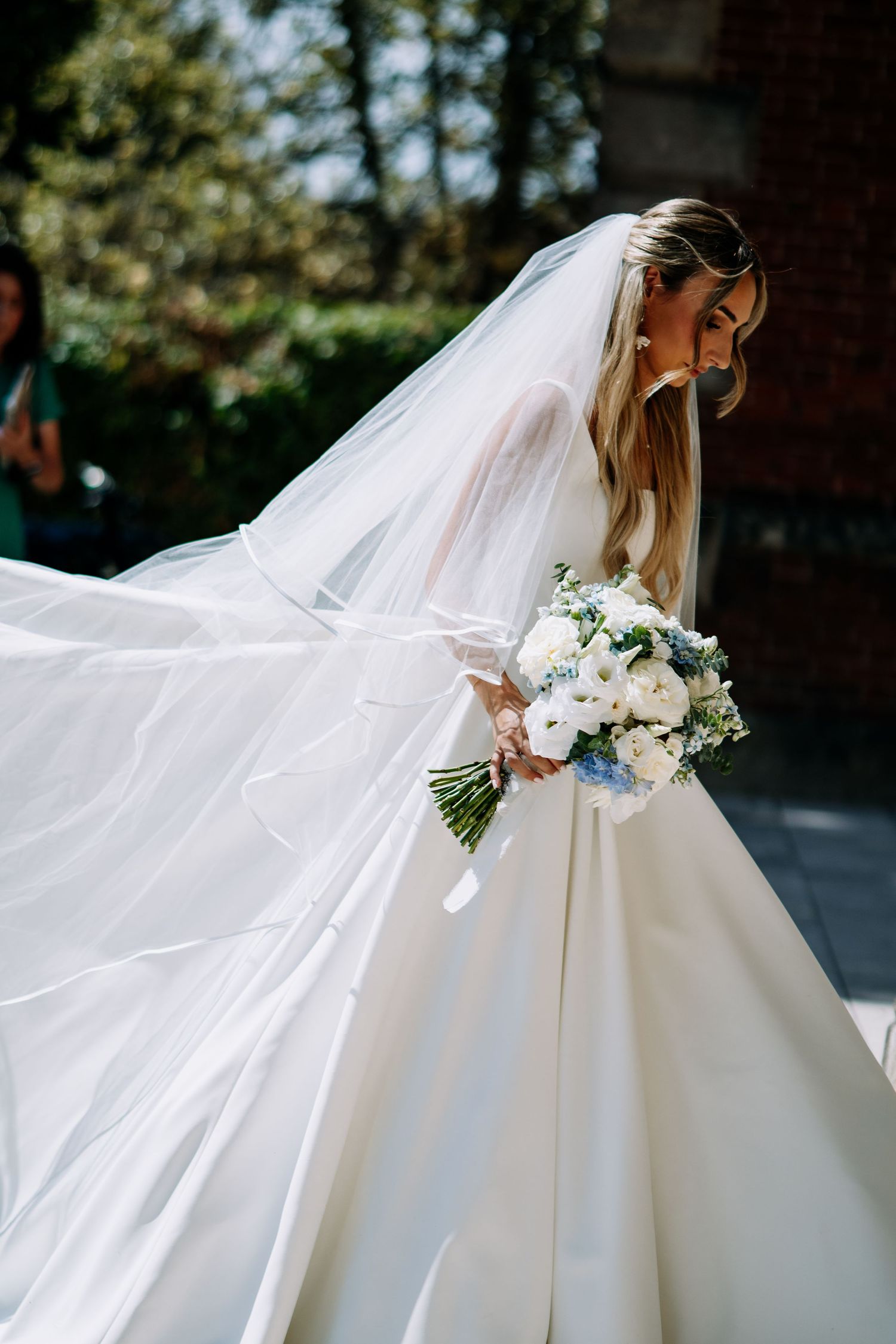 Bride in white satin ball gown with dramatic flowing veil holding white floral bouquet.