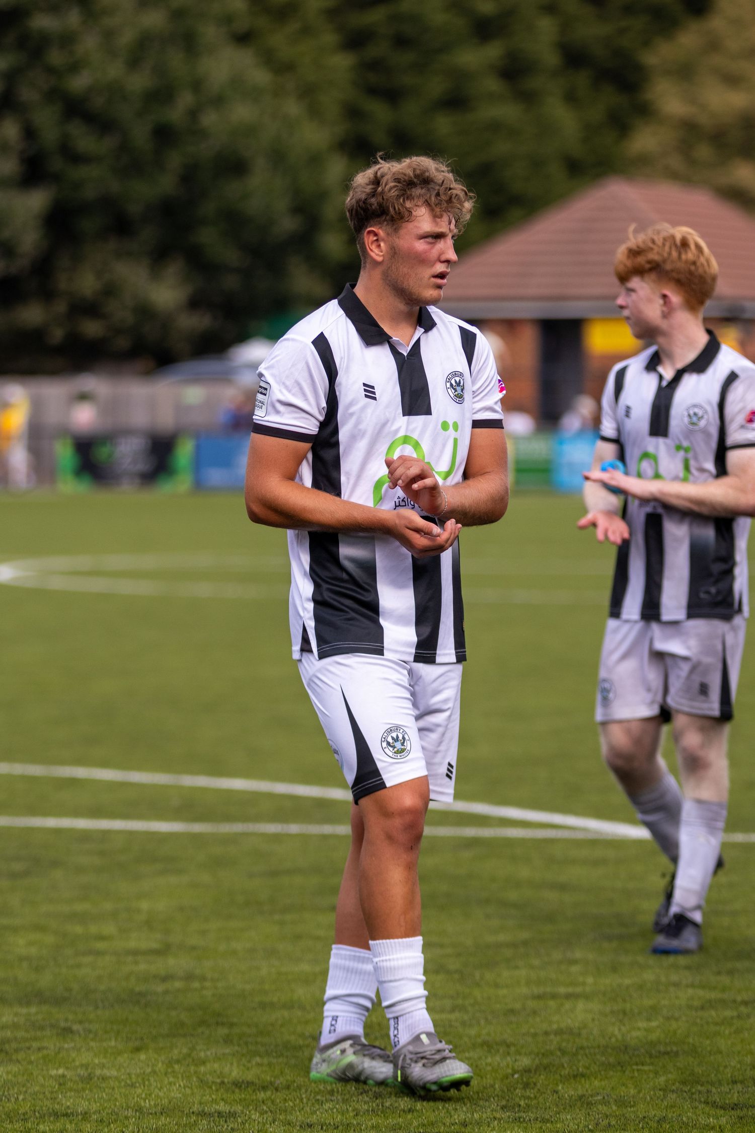 A young curly-haired footballer in black and white striped kit walks on the pitch after a match.