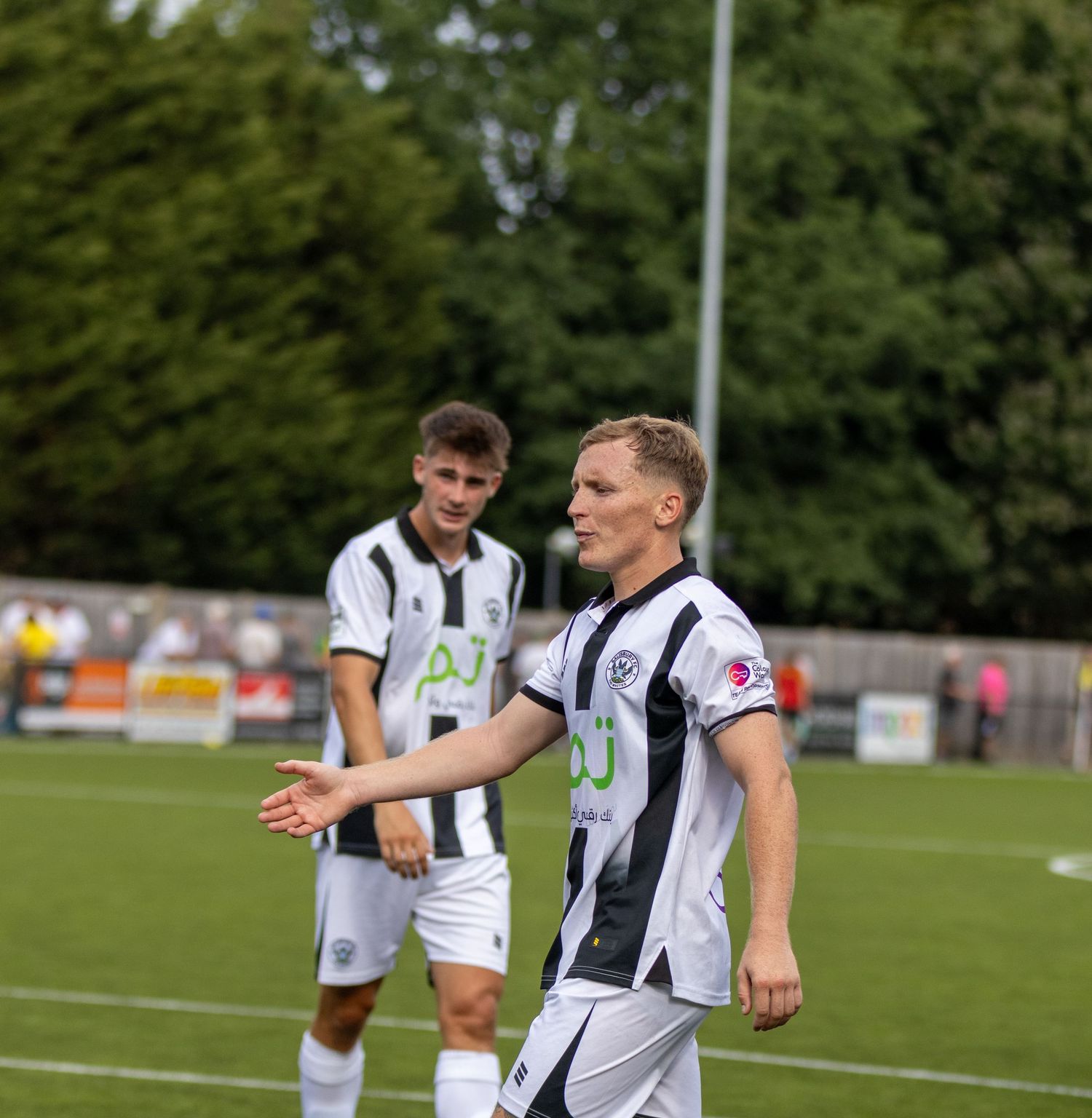 Two soccer players in black and white striped kits stand on the pitch during a football match.