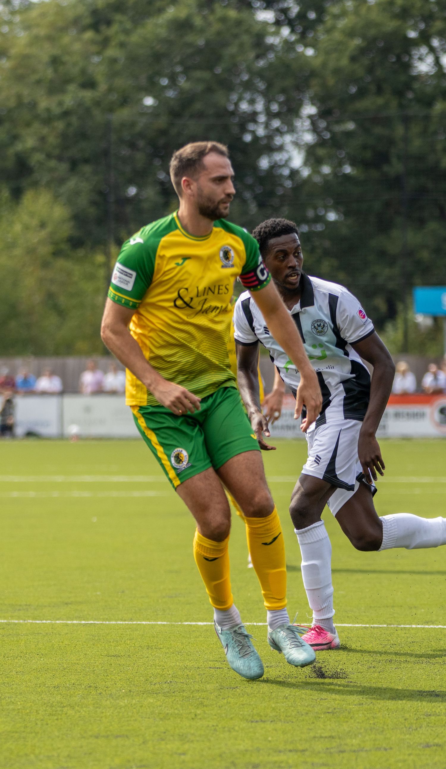 A footballer in yellow and green kit runs during a match against a player in black and white stripes on a grass pitch.