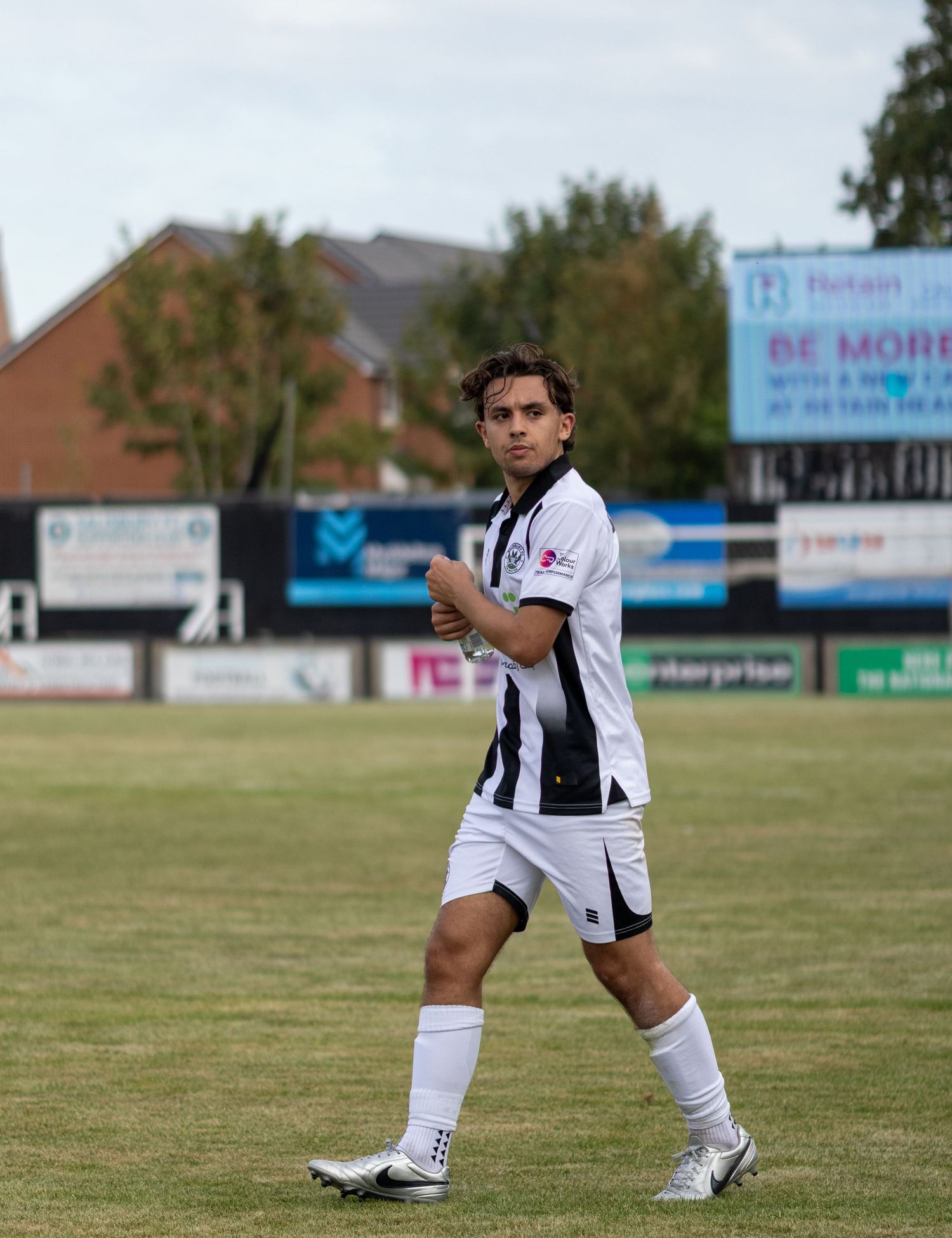 Male footballer in black and white striped kit gives thumbs up while walking on grass pitch.