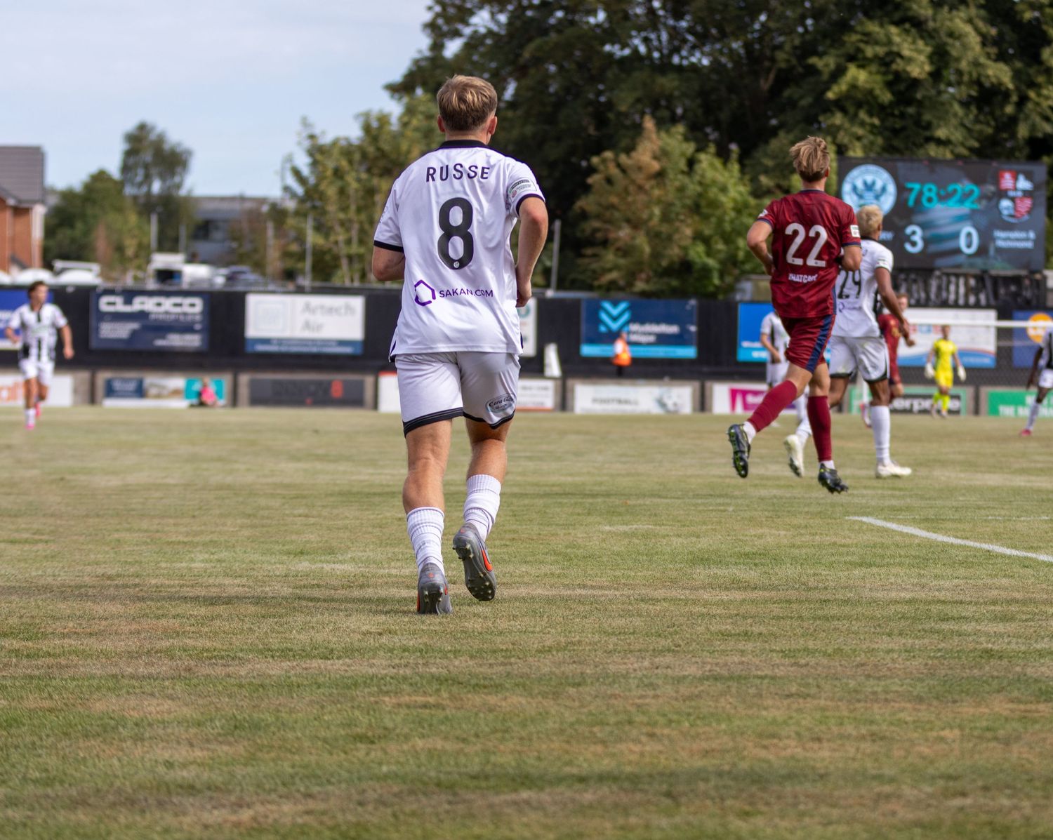 Player number 8 ROGGE walks on a sunny football pitch during a match with score 3-0 on the scoreboard.