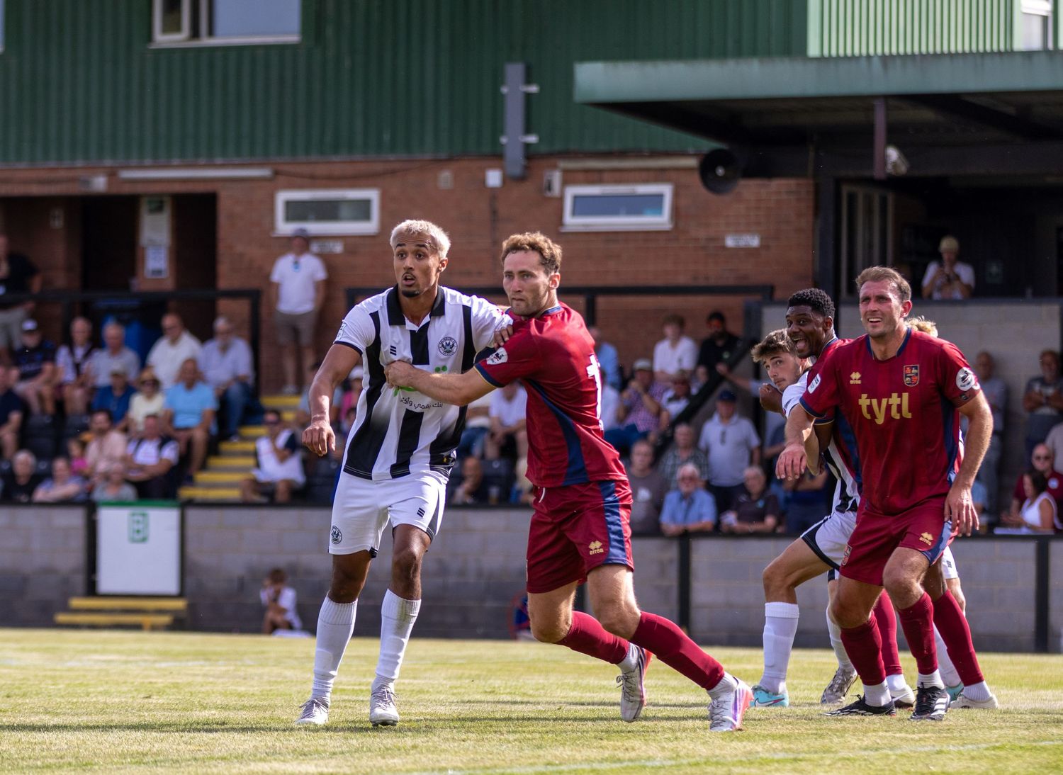 Footballers in black-white and red kits compete for position during a sunny non-league match.