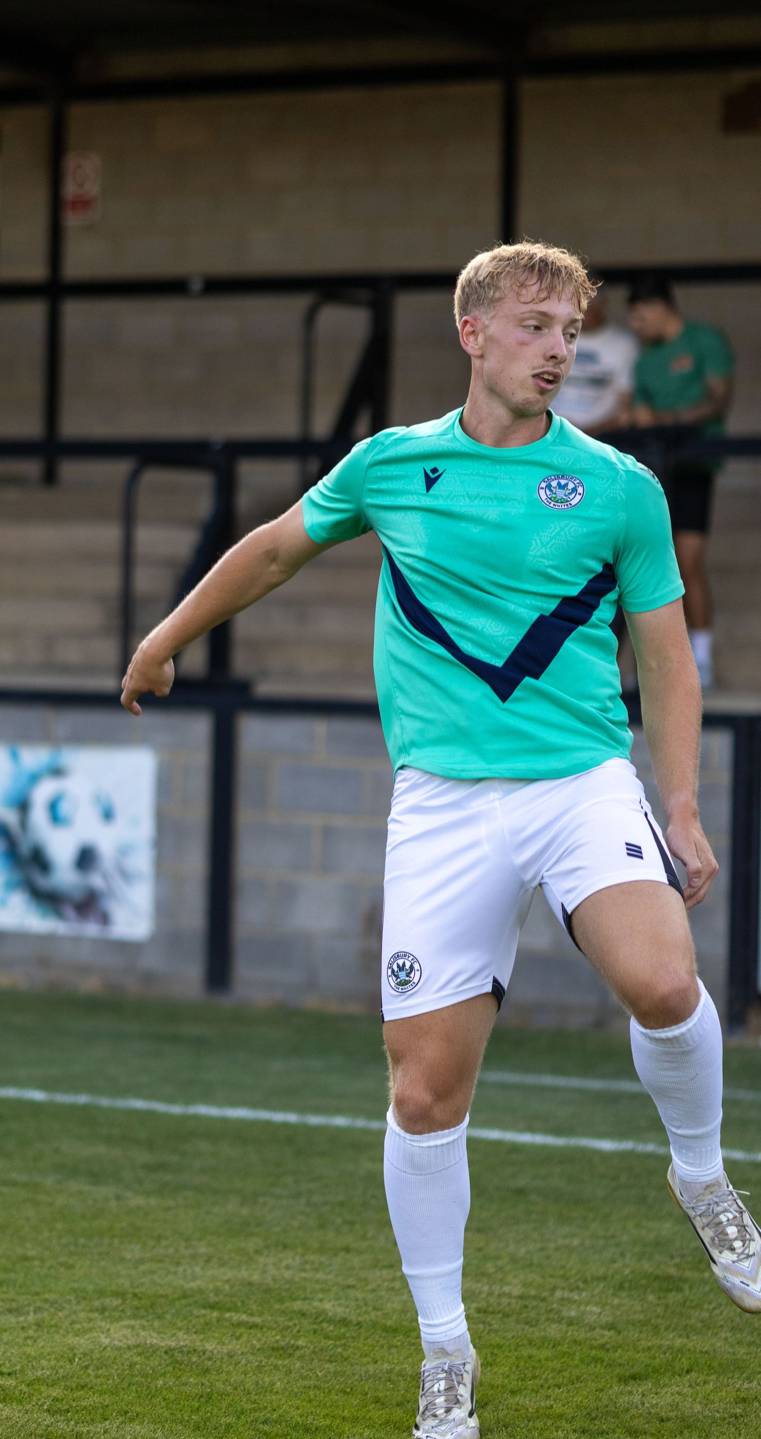 A young blonde soccer player in a teal green kit controls the ball during a football match.