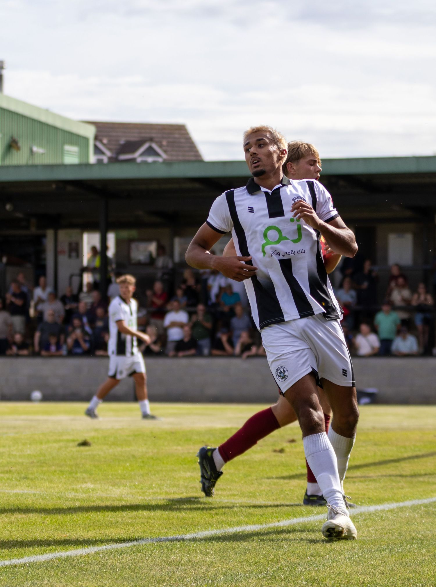 A soccer player in a black and white striped kit runs on the pitch during a sunny match.