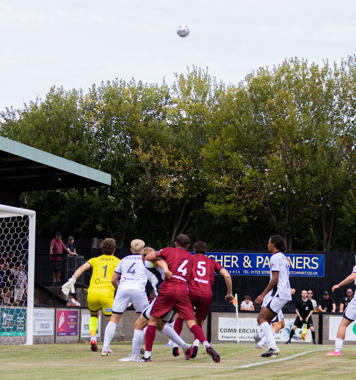 Players contest a high aerial ball near the goal during a football match on a cloudy outdoor grass pitch.