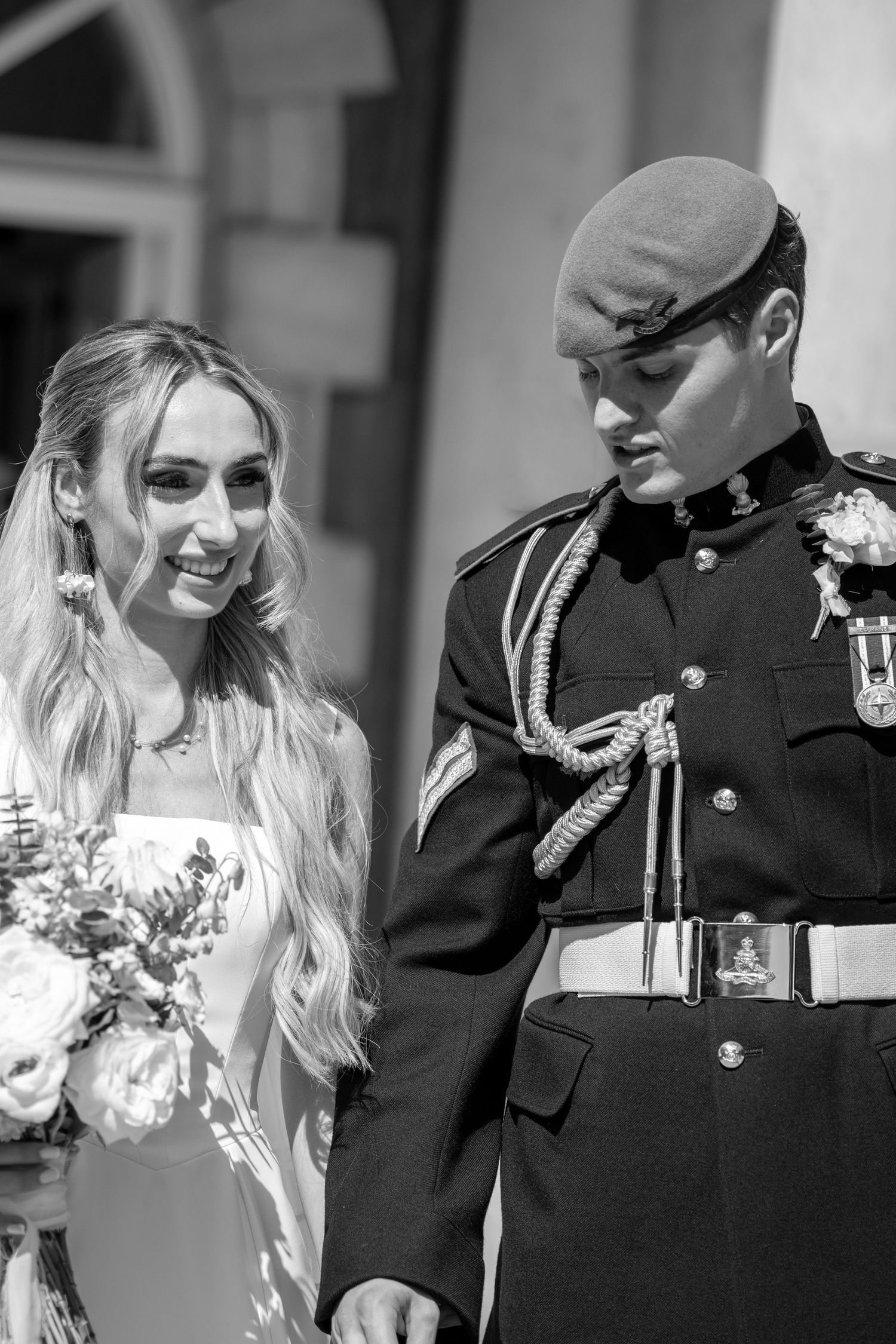 Black and white photo of smiling bride with long blonde hair and groom in military ceremonial uniform.