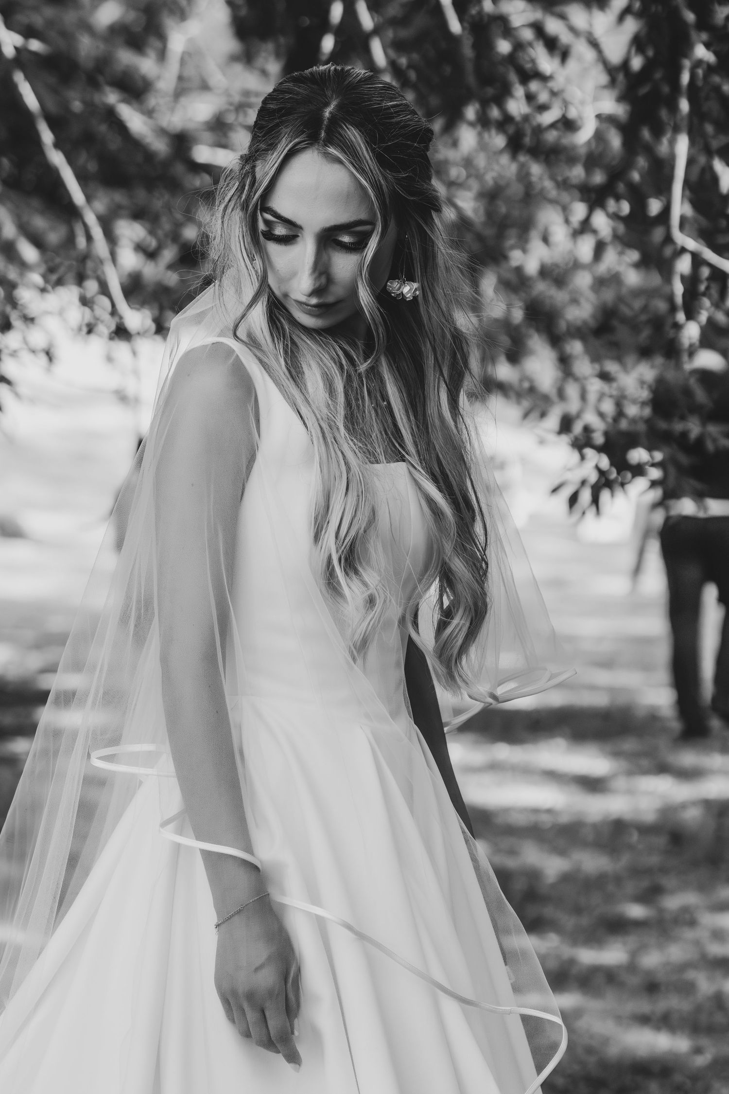 Black and white portrait of a bride in a white gown and veil among trees on her wedding day.