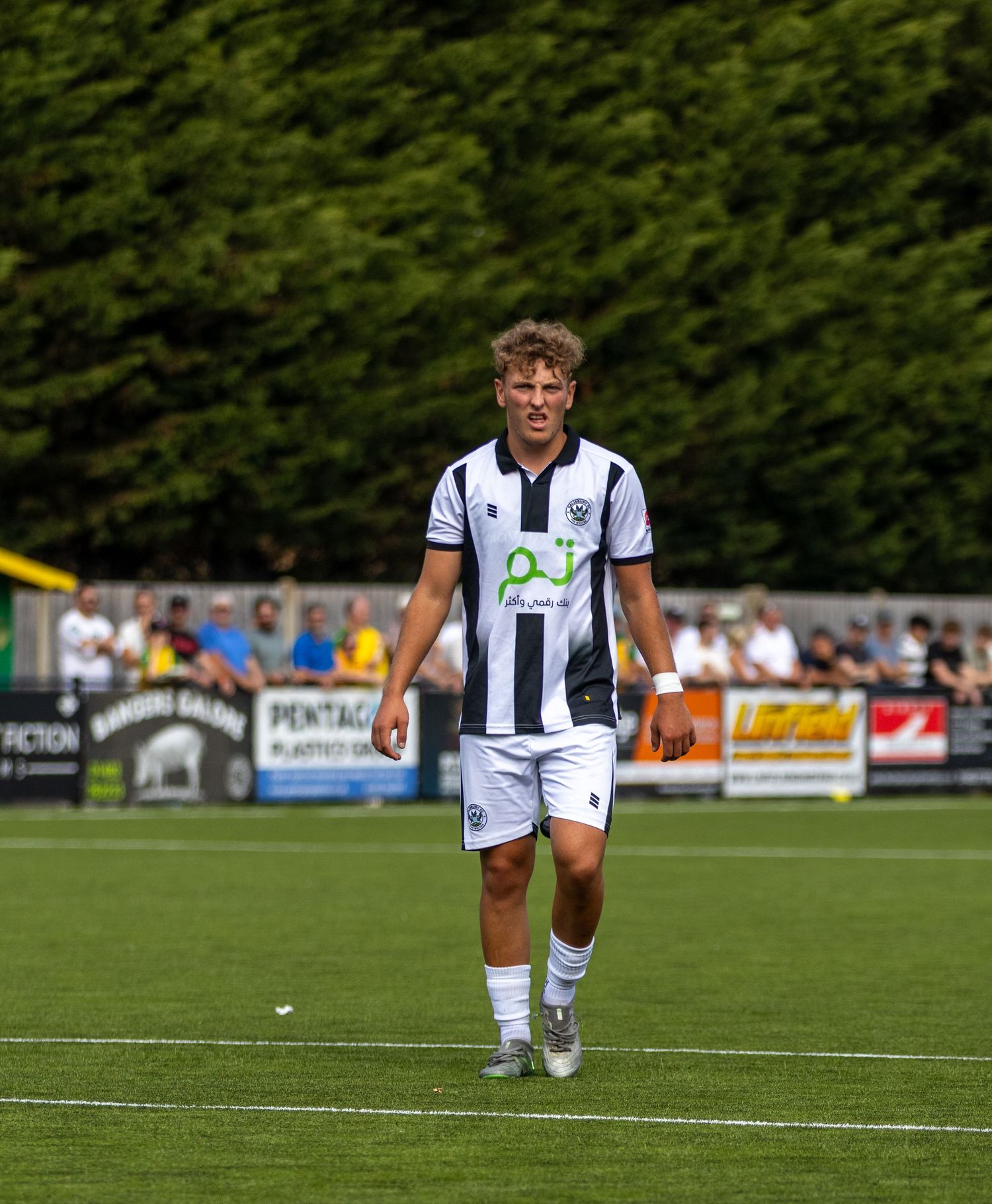 A blond footballer in white and black striped kit walks across a well-maintained football pitch.