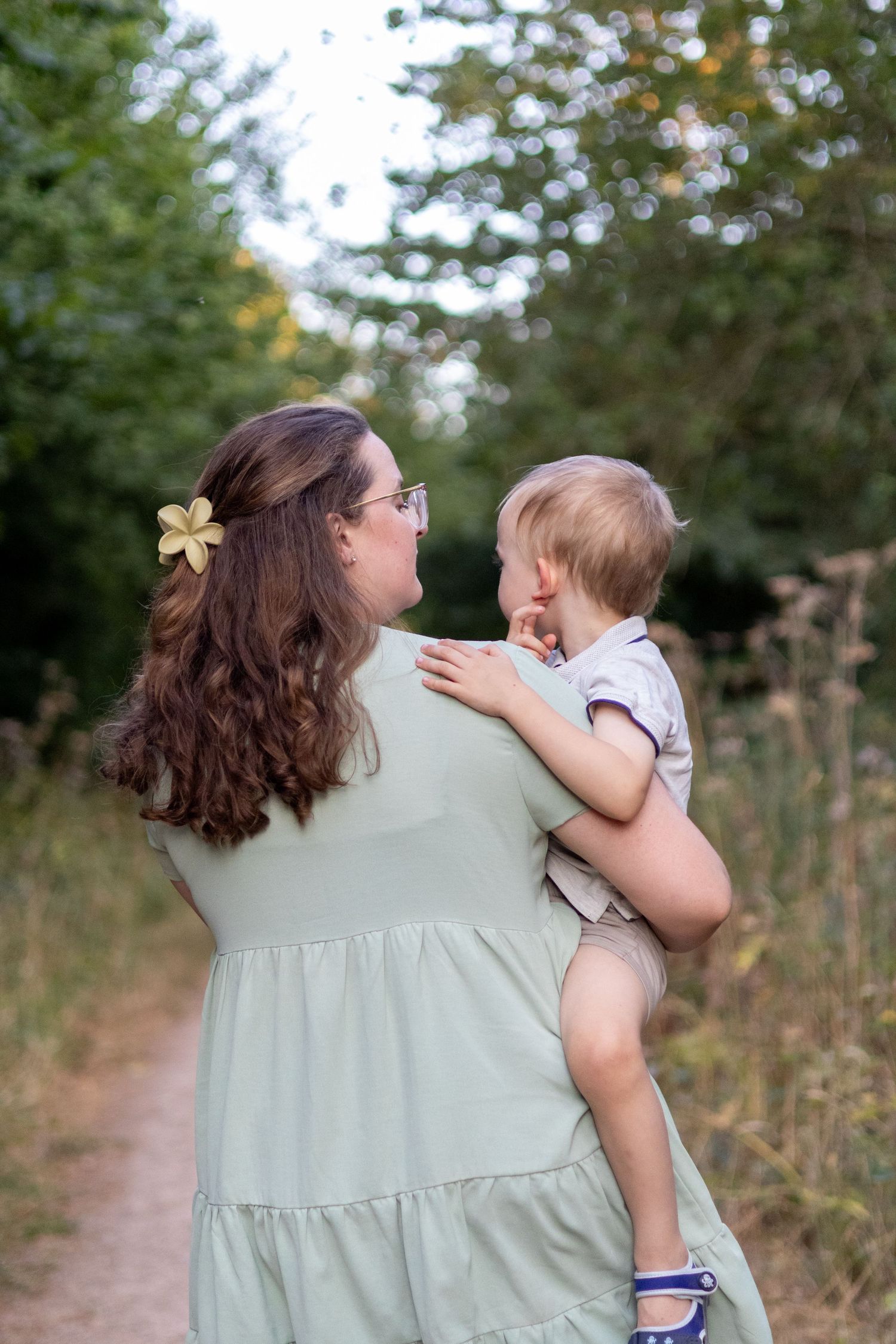 A mother with curly brown hair and flower clip holds her baby on a scenic green nature trail.