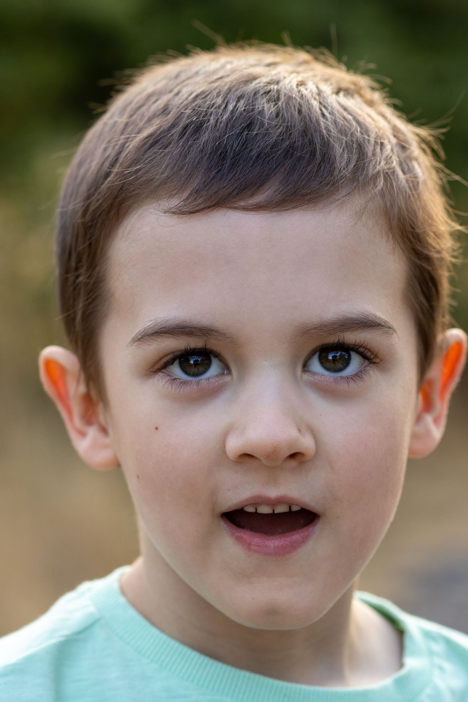 Close-up portrait of a young boy with brown eyes and short hair wearing a green shirt outdoors.