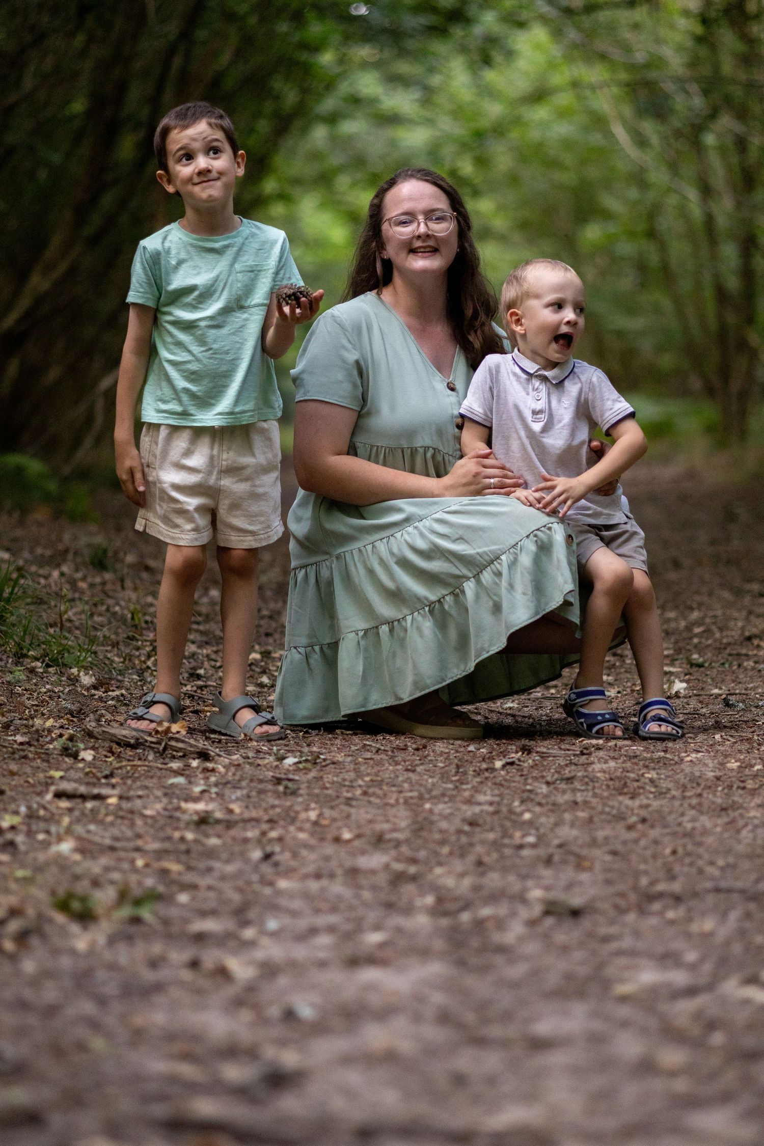 A smiling mother kneels between her two young sons on a leafy forest path.