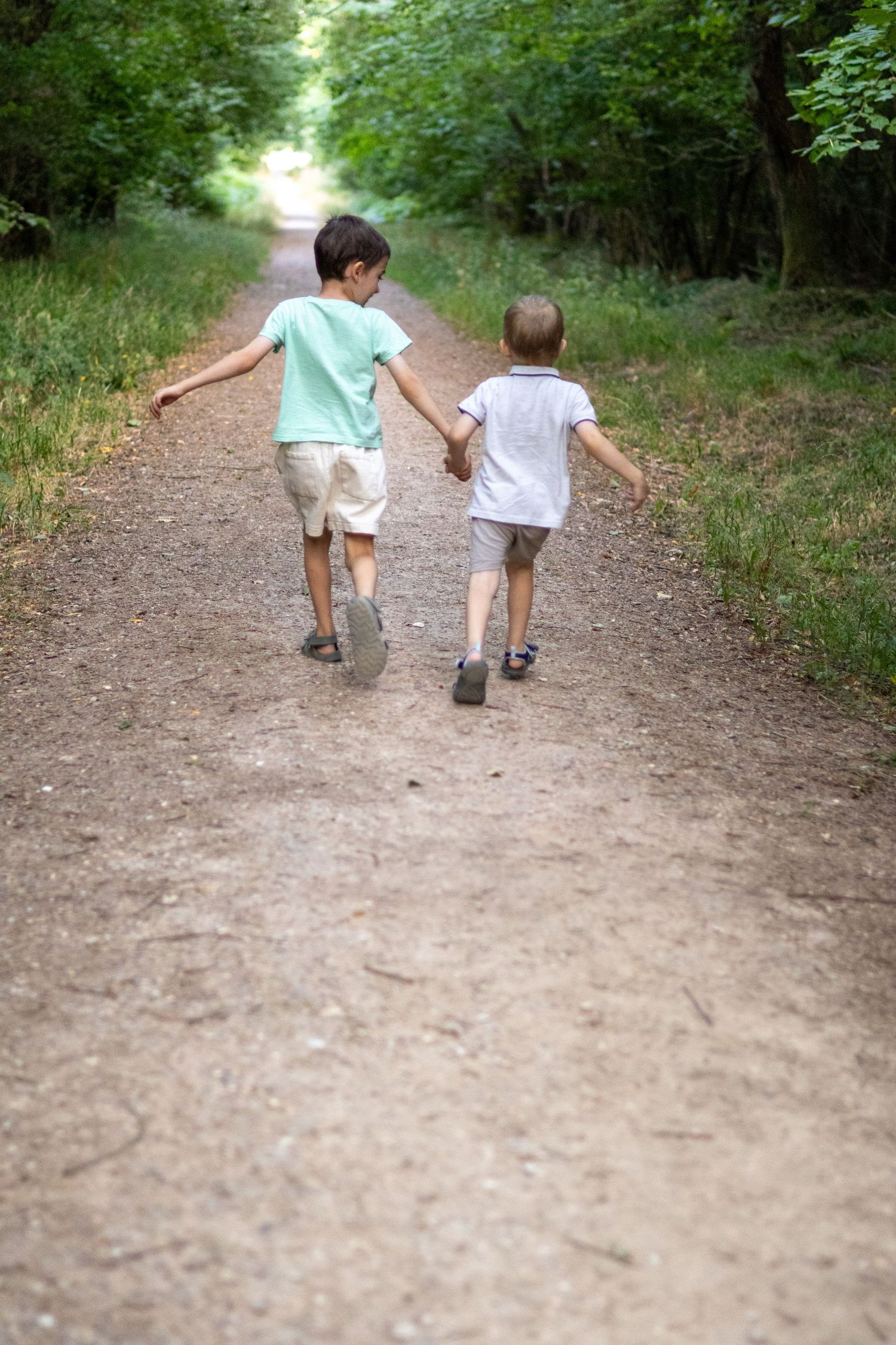 Rear view of two children holding hands running along a dirt path through green forest.