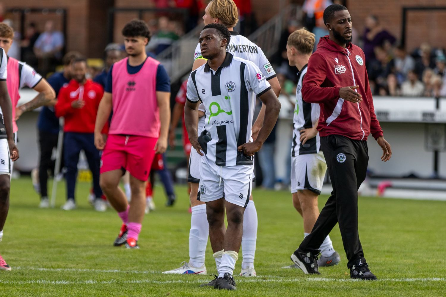 Footballers in black and white kit walk off pitch after match with coaching staff.