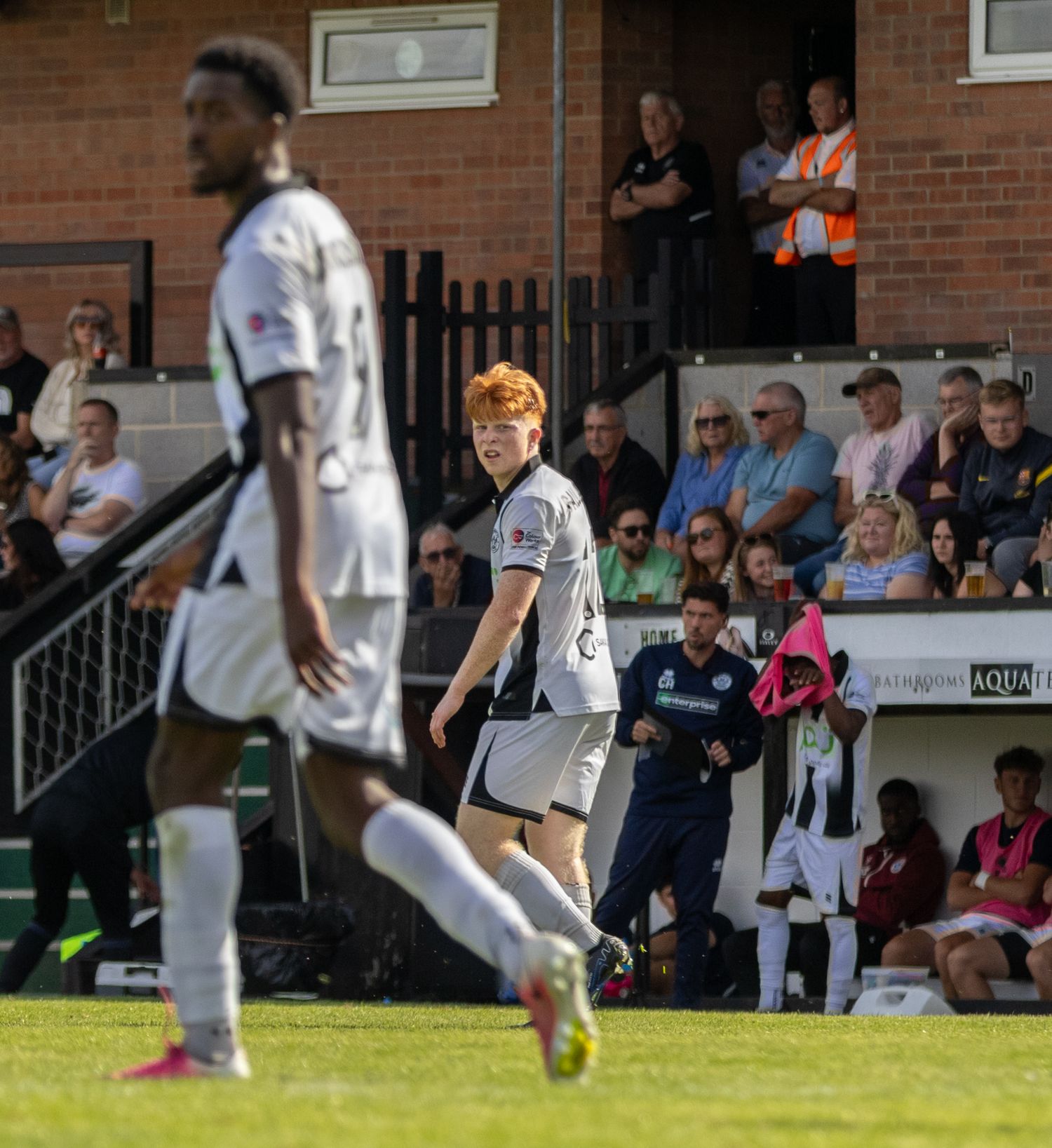 Red-haired footballer in grey kit walks on pitch watched by crowd at football stadium.
