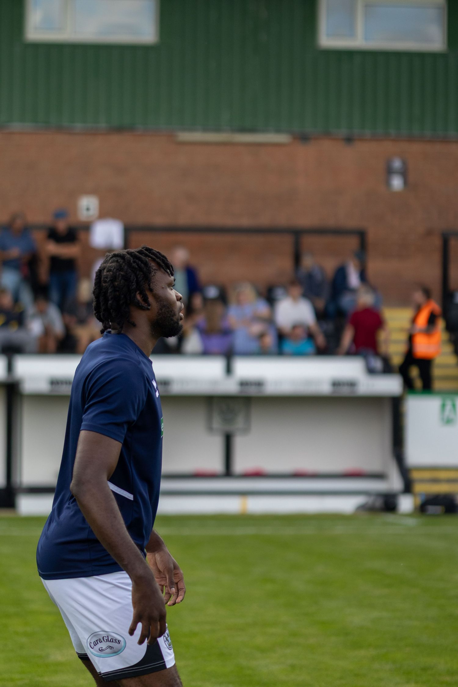 A Black male footballer with dreadlocks wearing a navy blue shirt and white shorts stands on a football pitch.