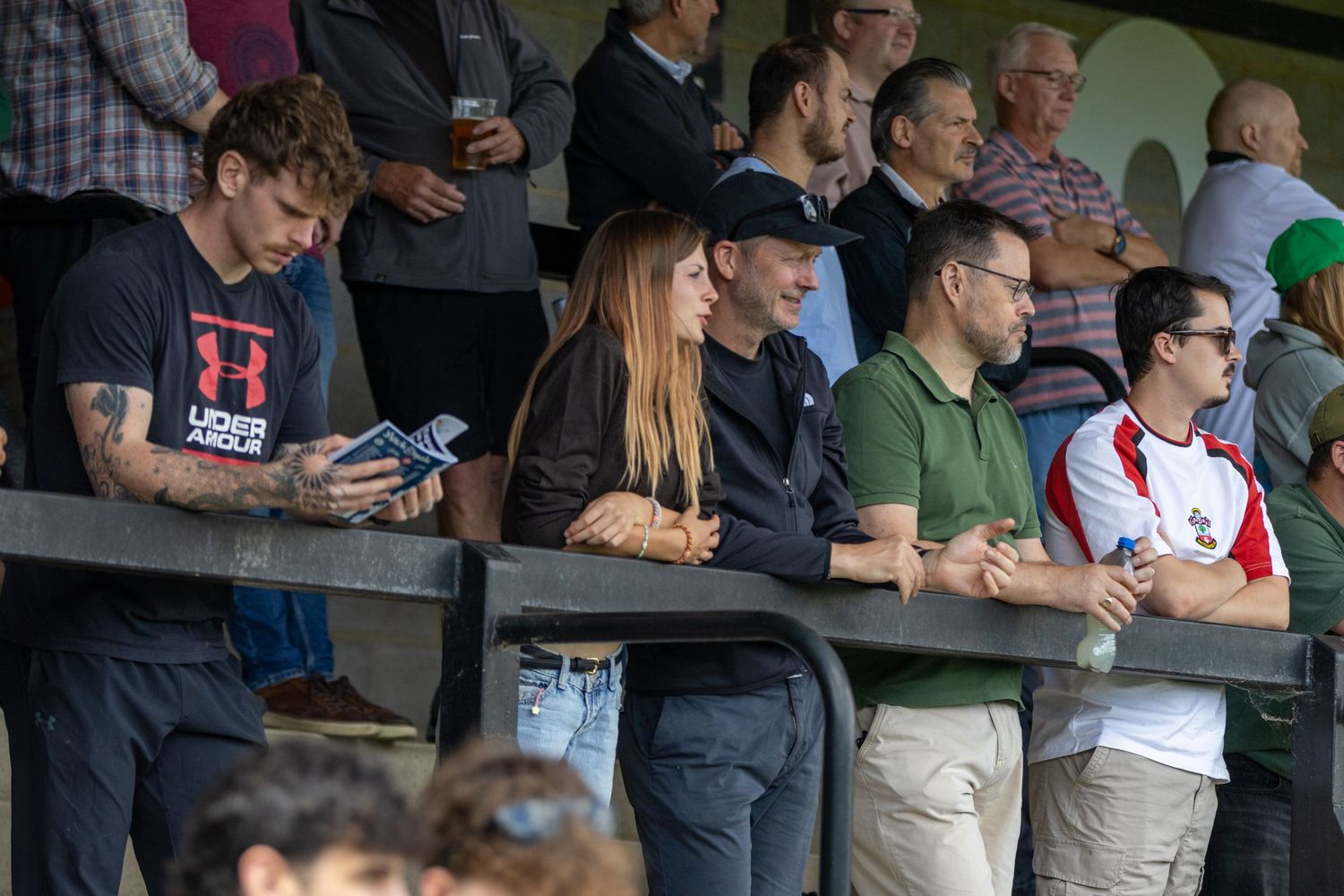 Football spectators watching a match from stadium stands including a tattooed man checking his phone.
