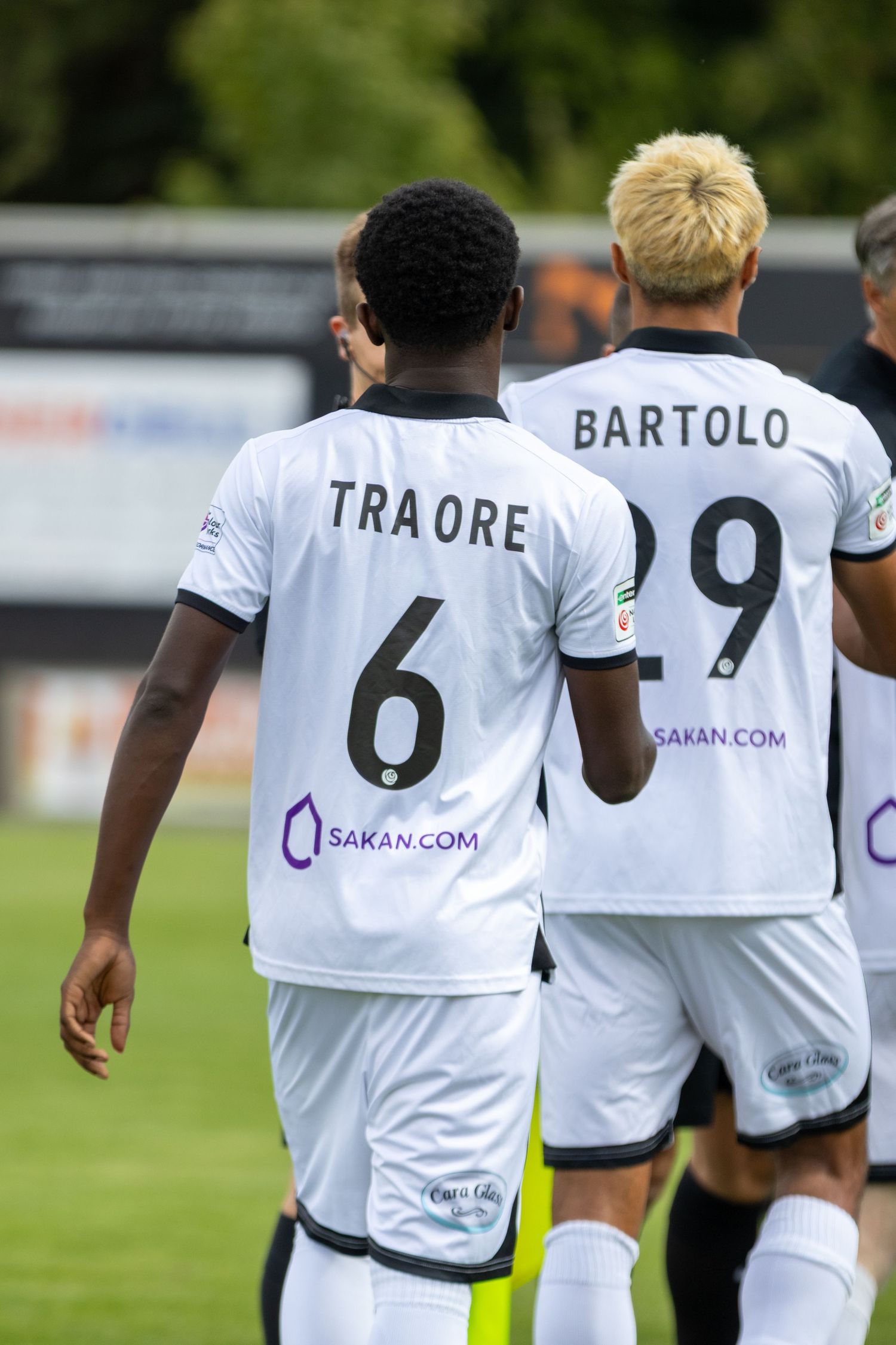 Traore wearing number 6 and Bartolo number 29 in white Sakan.com sponsored football kits walking.