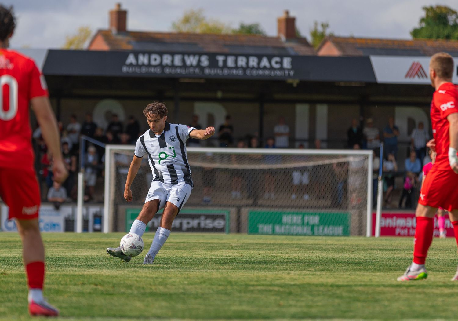 A footballer in black and white kit strikes the ball during a match at Andrews Terrace ground on a sunny day.