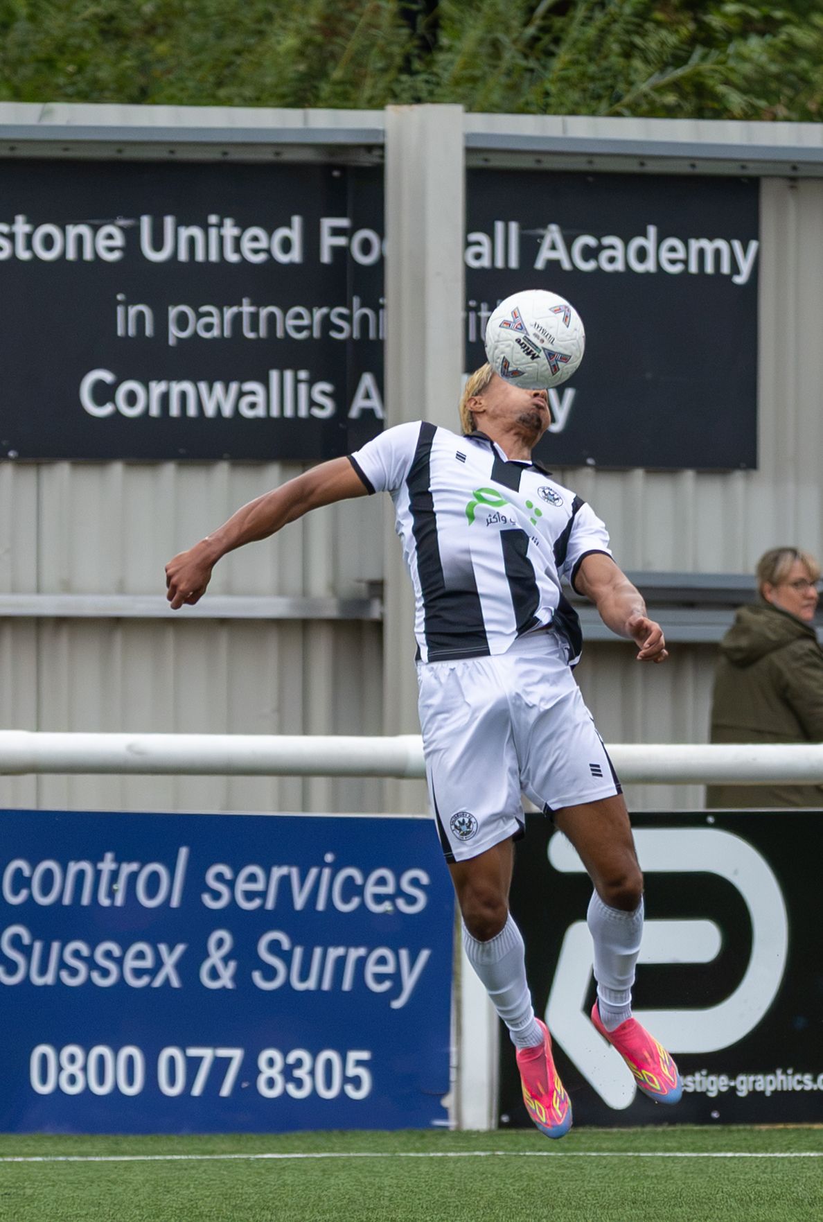 A soccer player in white and black kit heads the ball during a match at Maidstone United Football Academy.