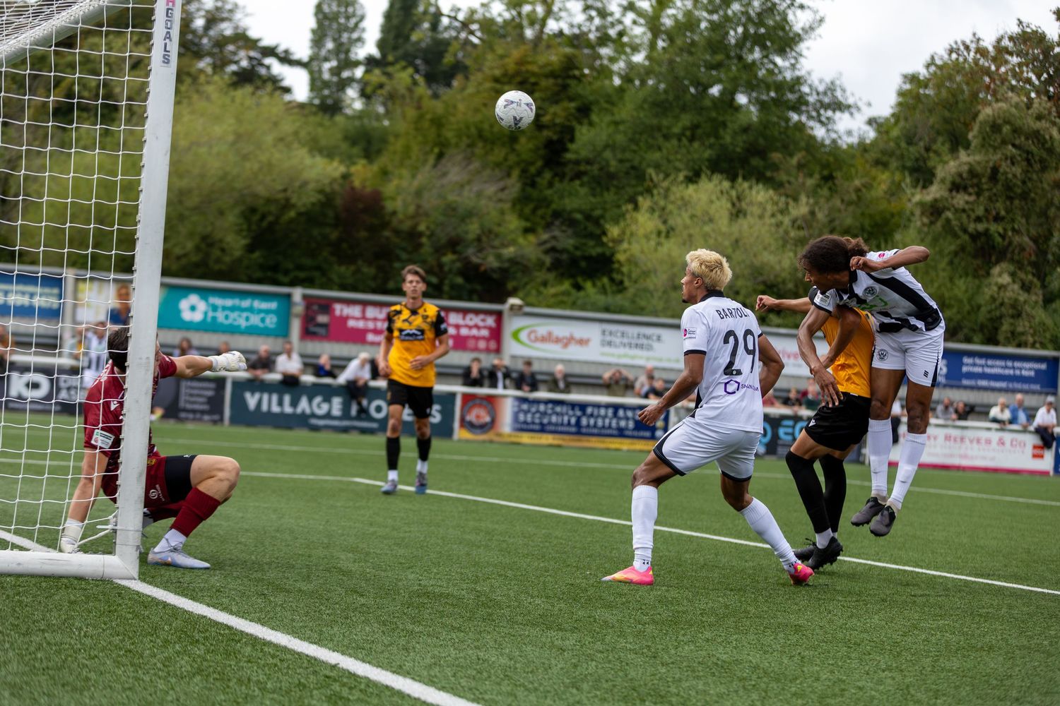 Soccer players compete for a header near the goal during an outdoor match on a green pitch.