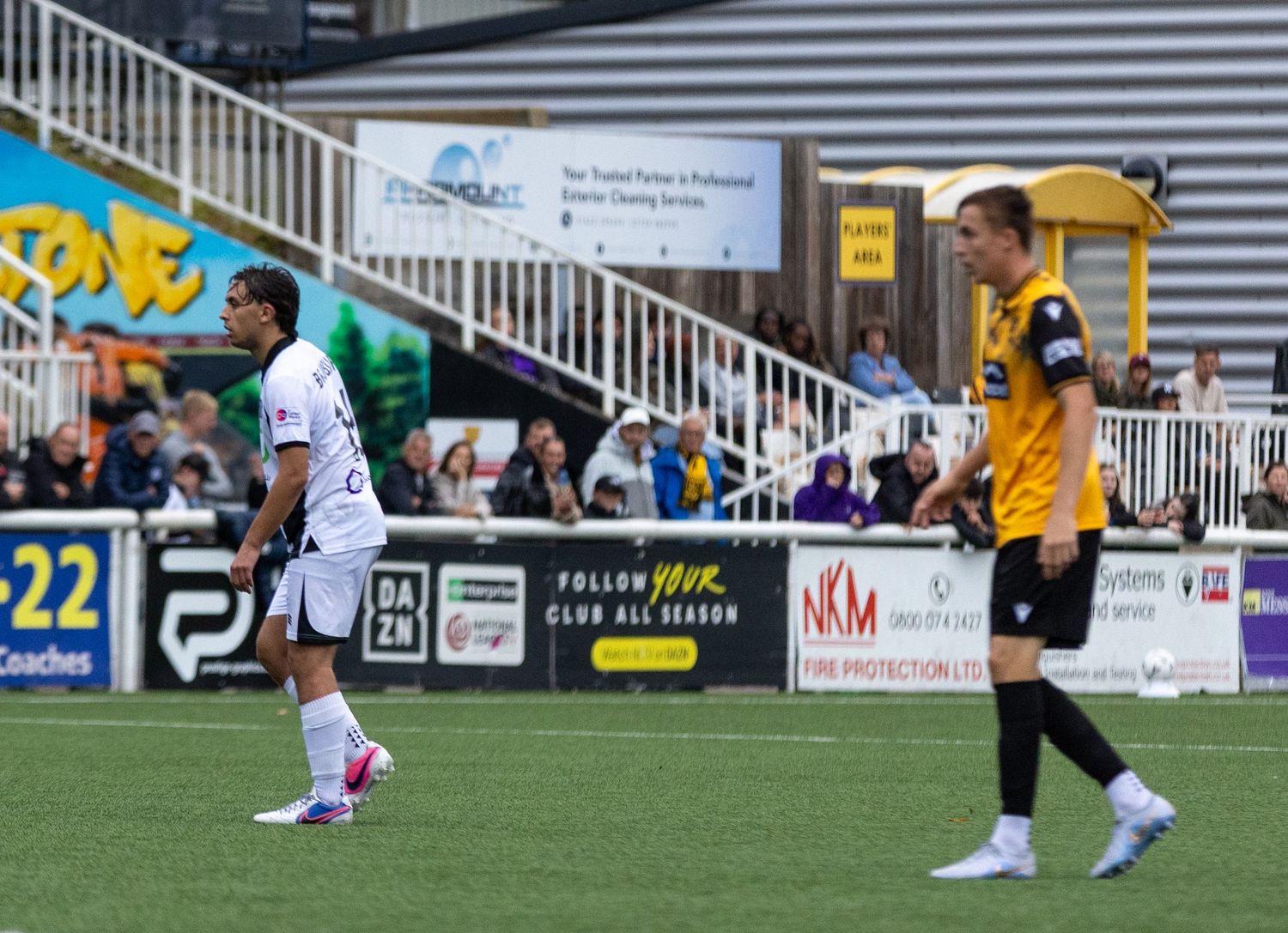 Two soccer players on a green pitch during a match, one in white and one in yellow, with fans and advertising boards visible.