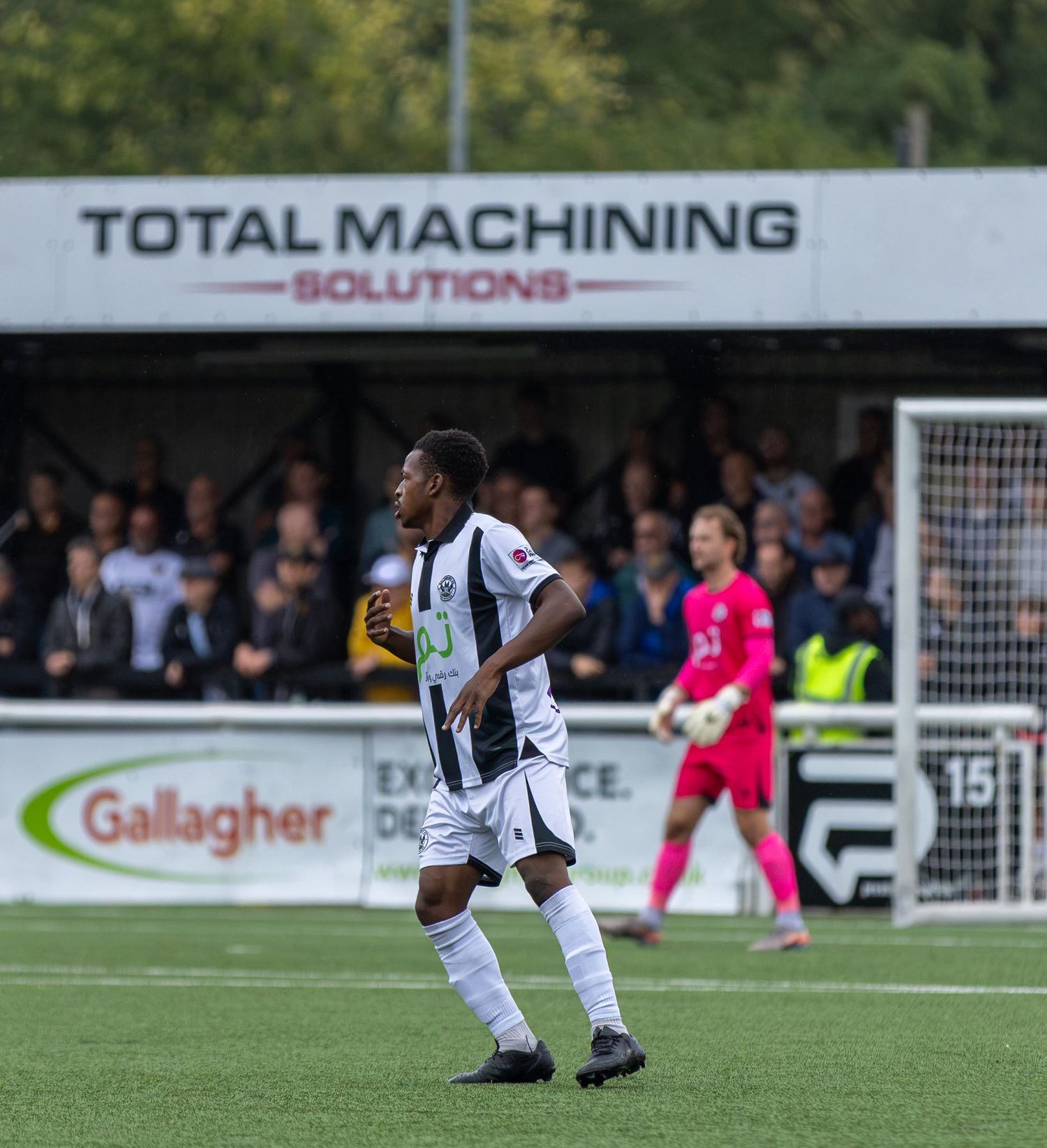 A footballer in black and white kit plays on a pitch in front of a Total Machining Solutions advertising board.