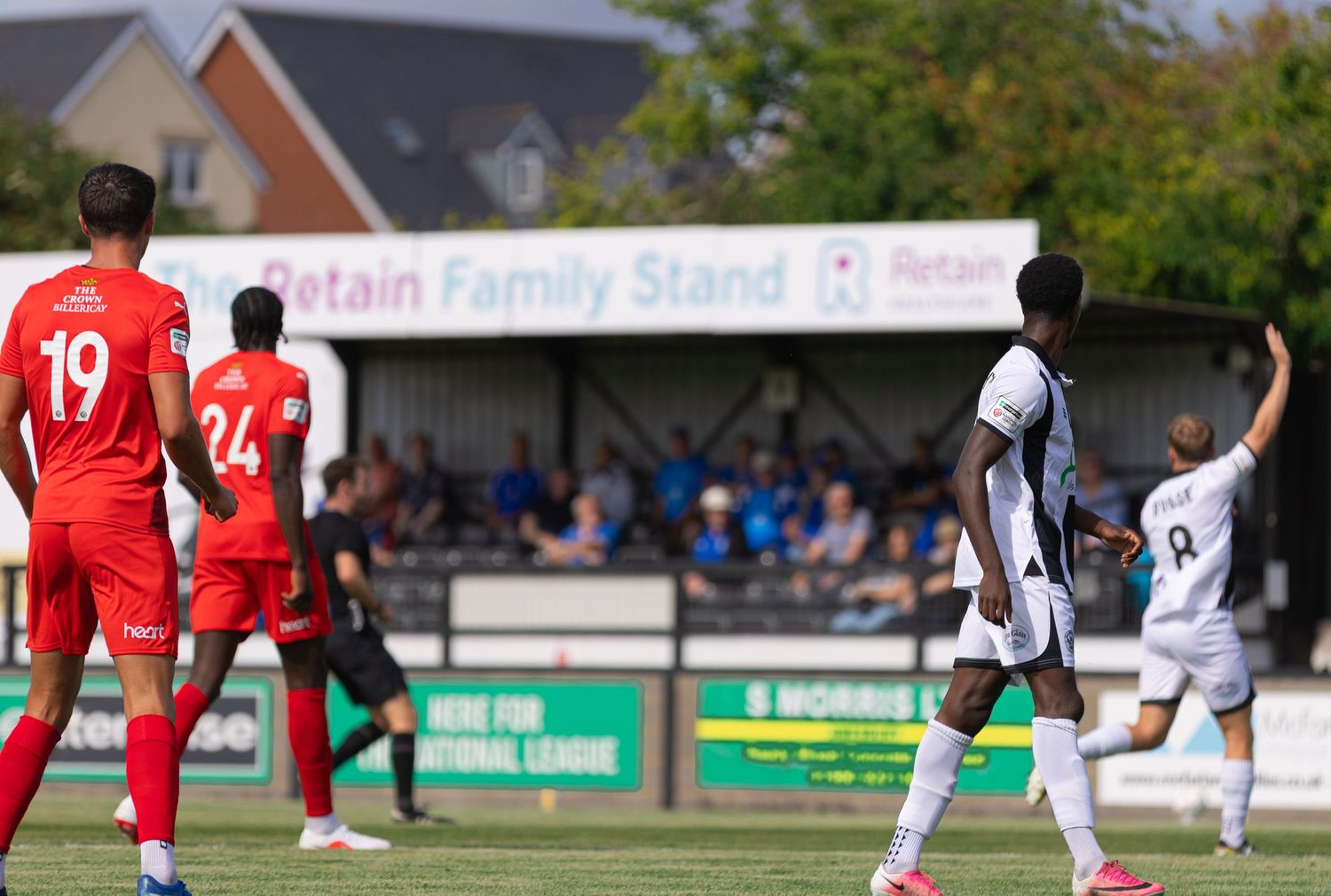 Soccer players in red and white uniforms compete on a grass field near the Retain Family Stand on a sunny day.