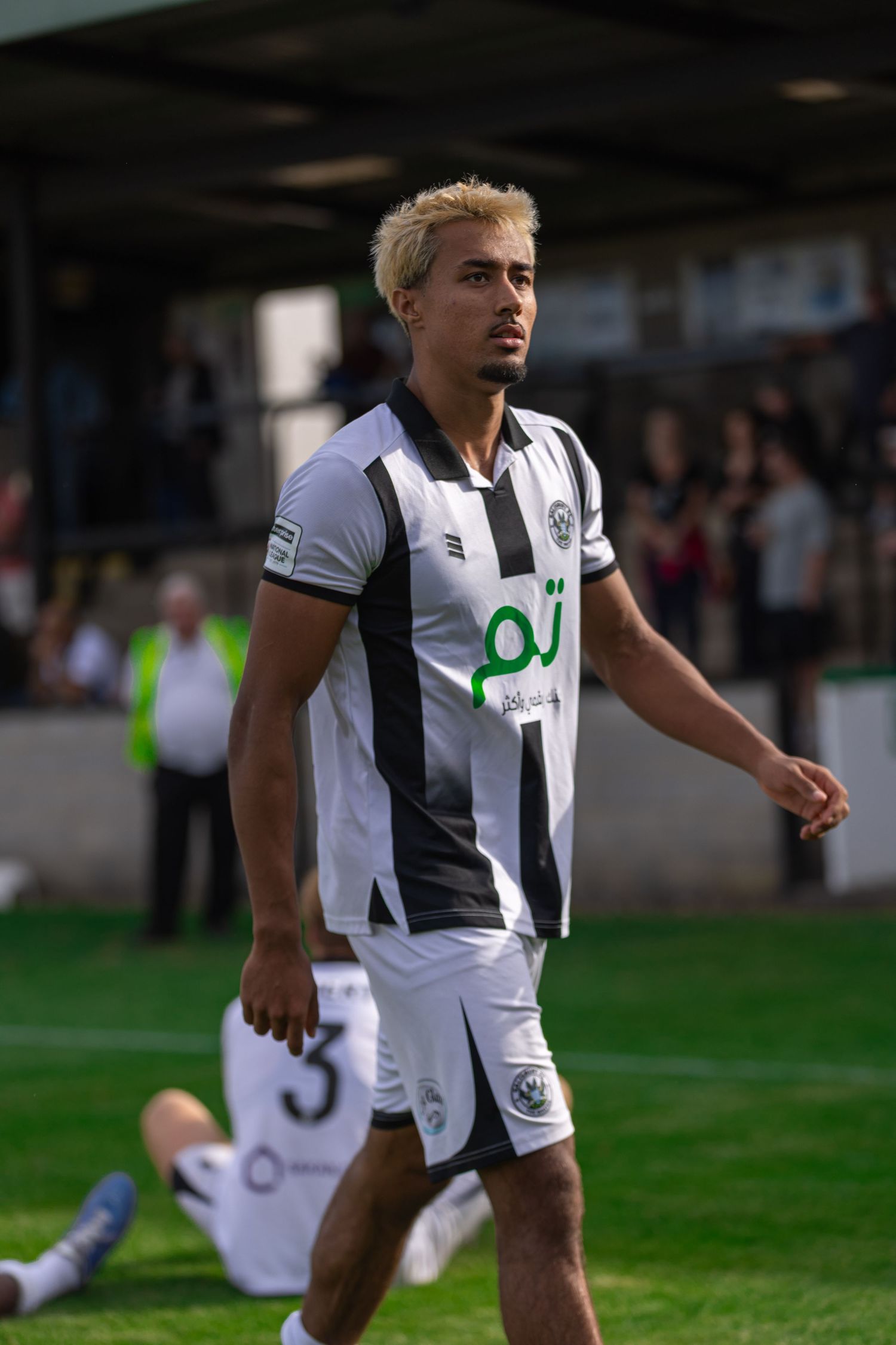 Soccer player in black and white striped kit with blonde hair walks on pitch during a match.