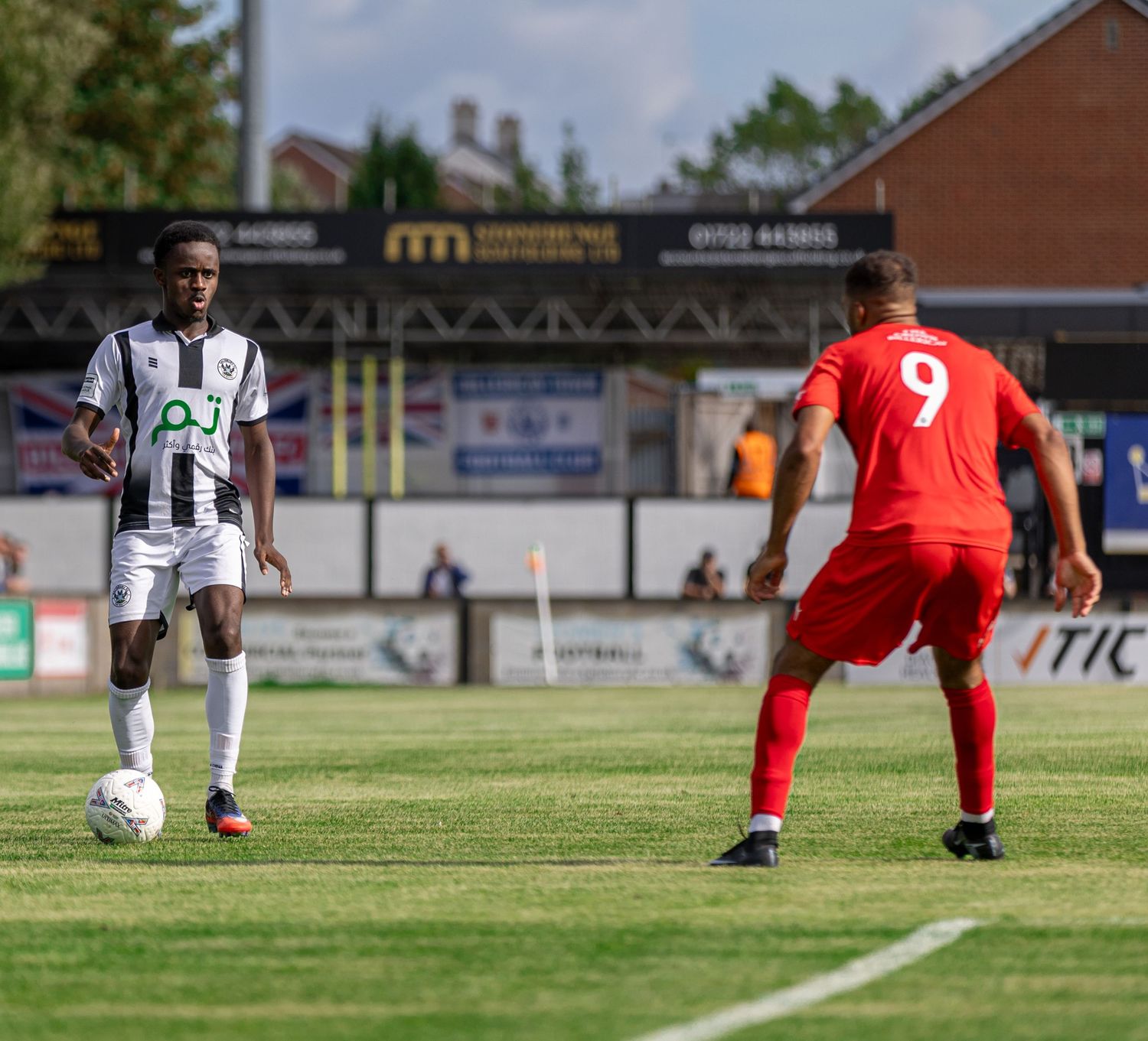 Two soccer players compete on a green pitch, one in black and white kit dribbling, another in red number 9 jersey defending.