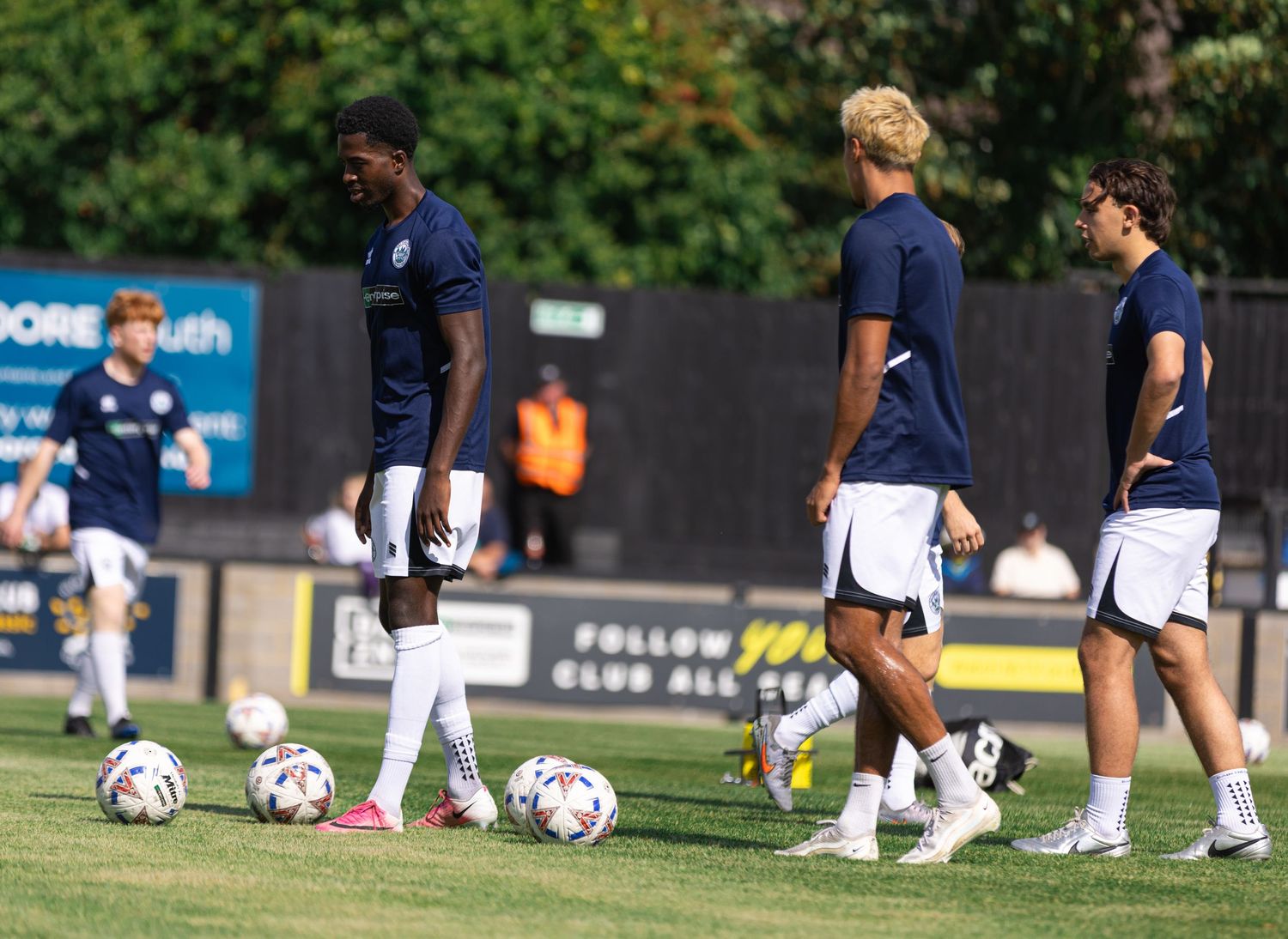 Soccer players in navy uniforms warm up on a green field with multiple balls during a sunny outdoor training session.