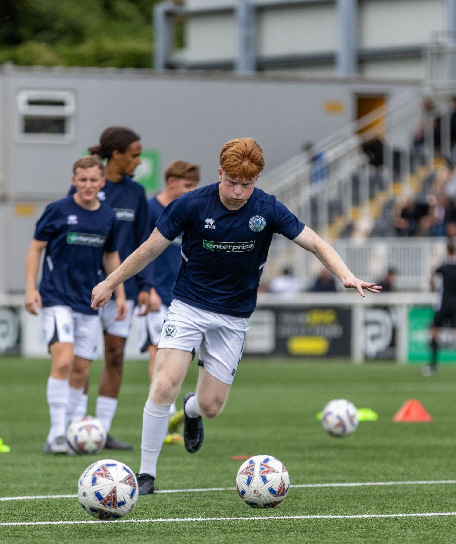Young red-haired footballer in navy kit dribbling a ball during a youth soccer training session on a green pitch.