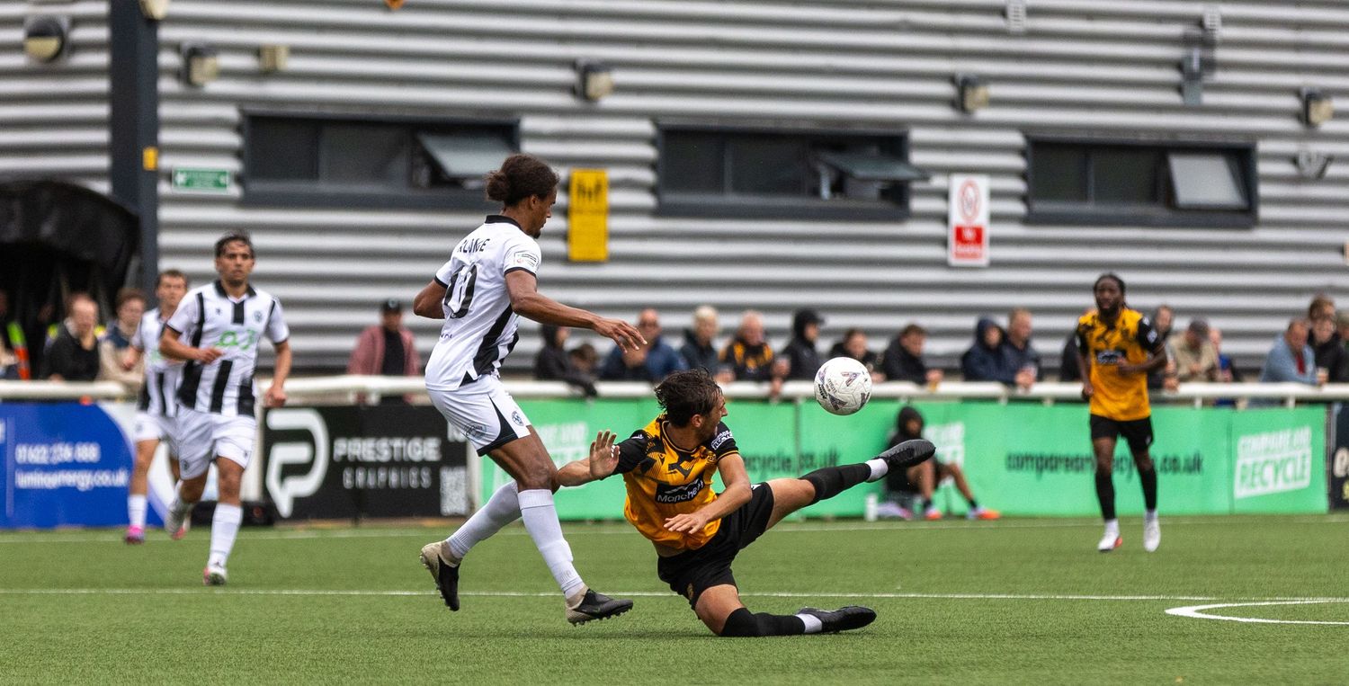 Soccer player in white kit shoots past sliding goalkeeper in yellow during an outdoor match on a green field.