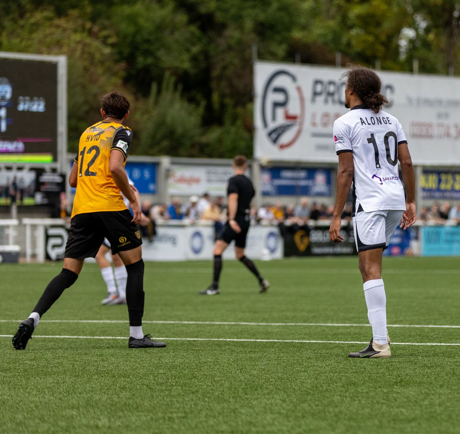 Two soccer players, numbers 12 and 10, compete on a green pitch during a sunny outdoor match.