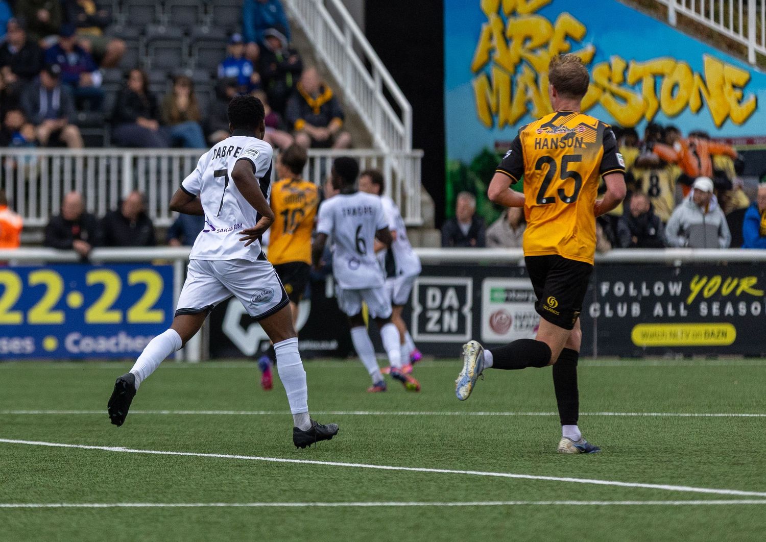 Soccer players in action during a match, with one player in white kit sprinting past a defender in yellow and black.